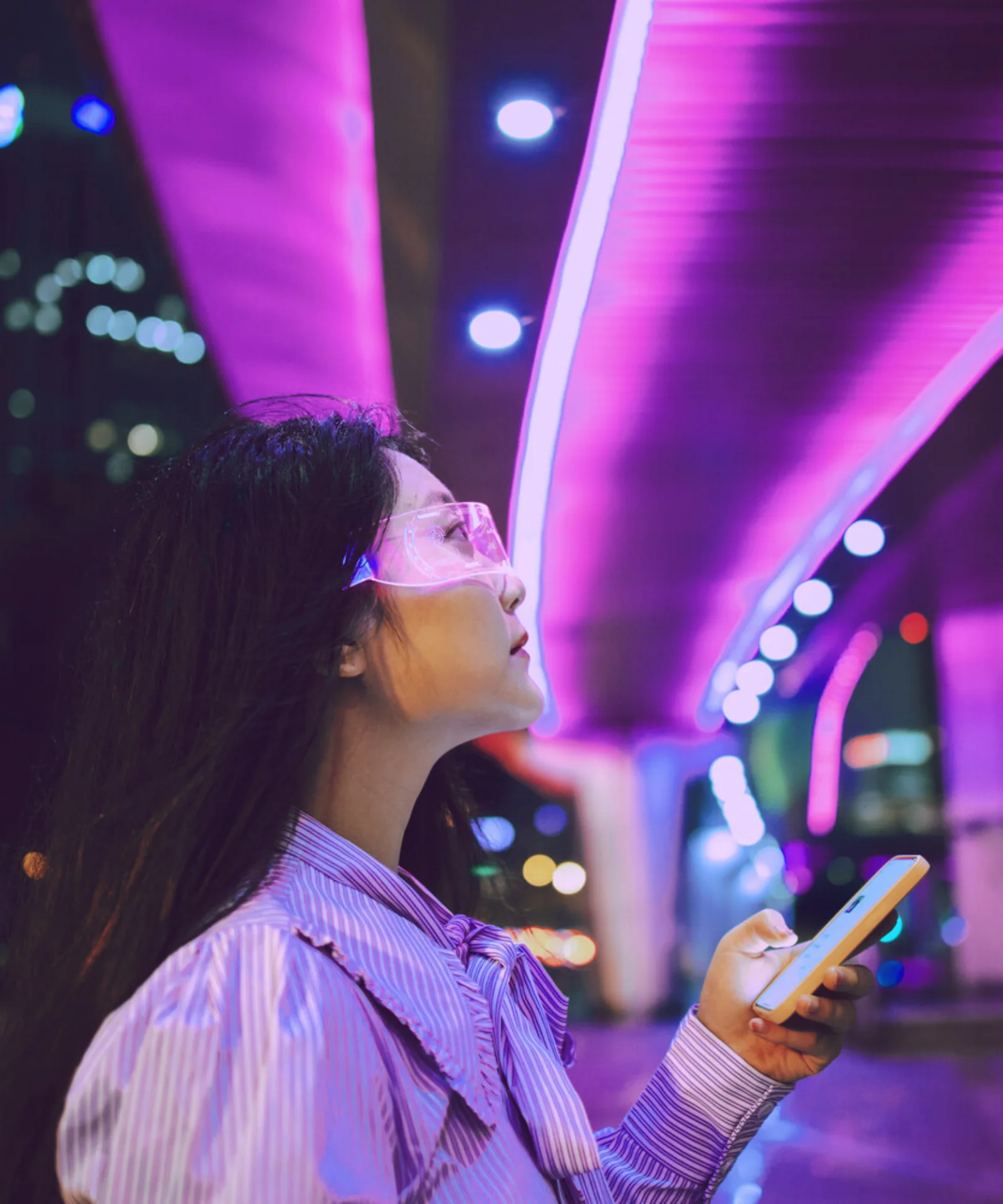 A woman holding a smartphone and looking up at vibrant neon lights on a modern urban bridge at night, symbolizing technology and urban life.