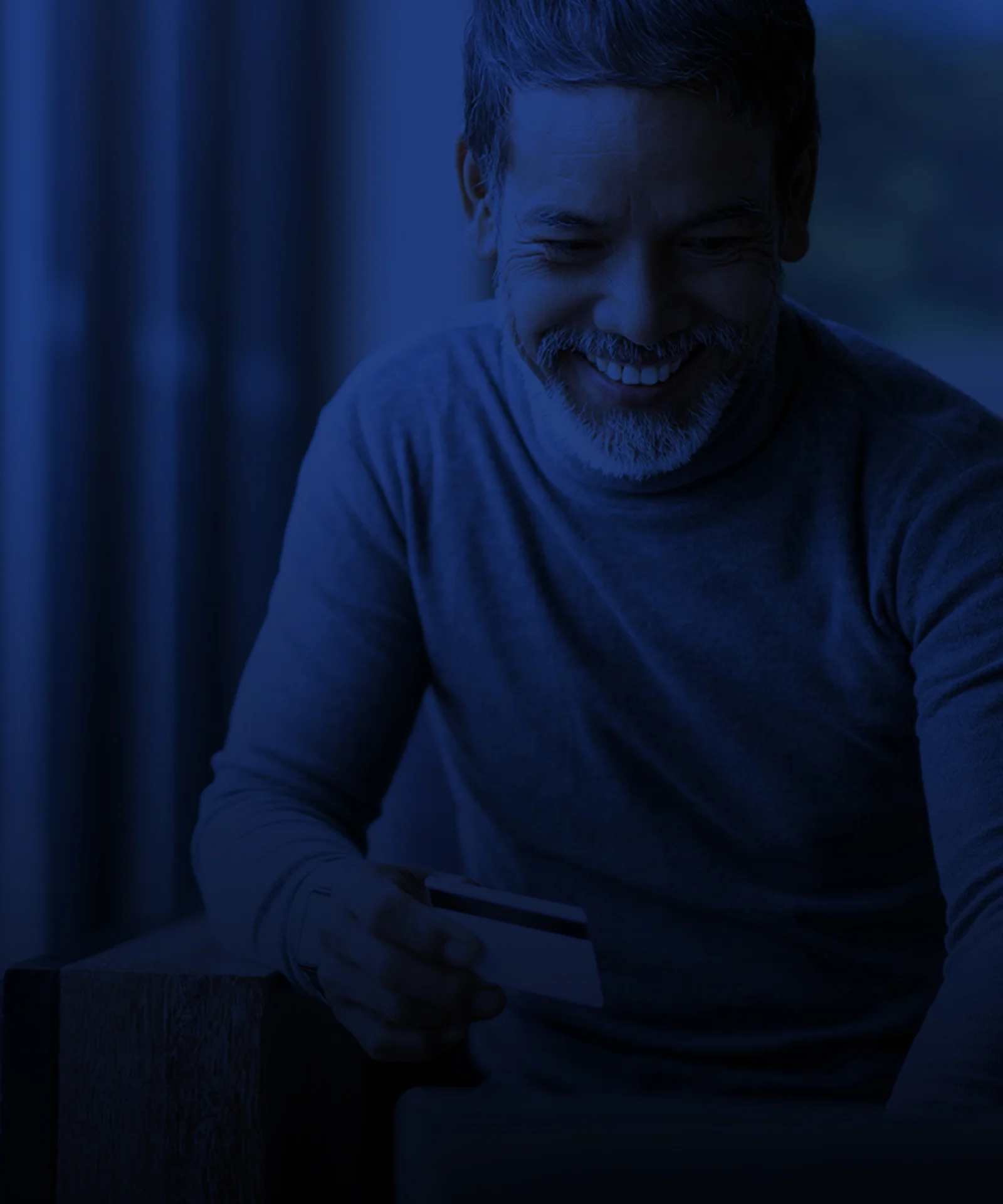 A man smiling while holding a credit card, making an online payment in a dimly lit, modern setting.