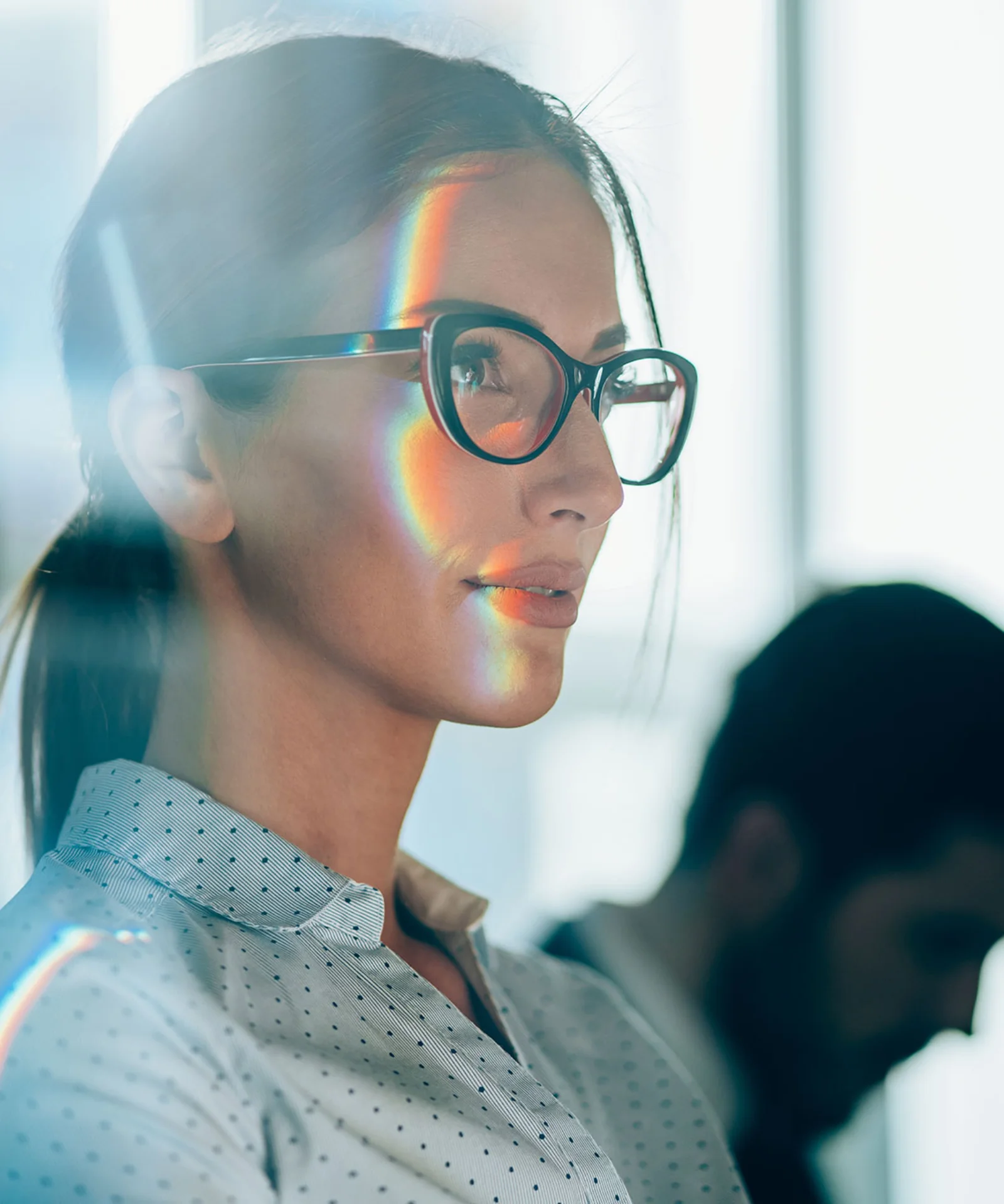 A confident woman wearing glasses looks ahead with a focused expression, with a subtle rainbow reflection on her face, symbolising strategic thinking and modern business acumen.