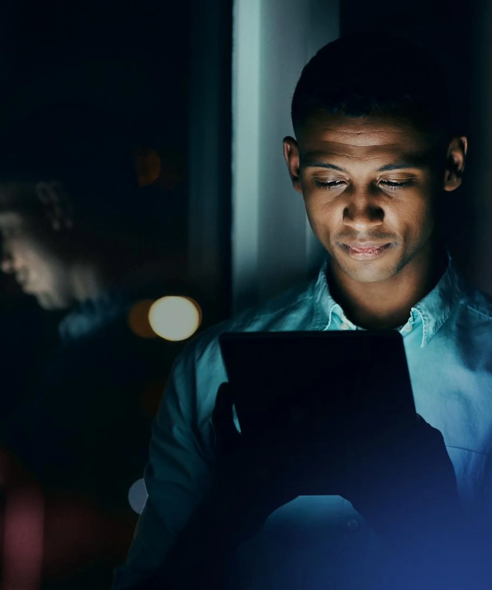 Man looking at a tablet screen at night, standing by a window with city lights reflecting on the glass.