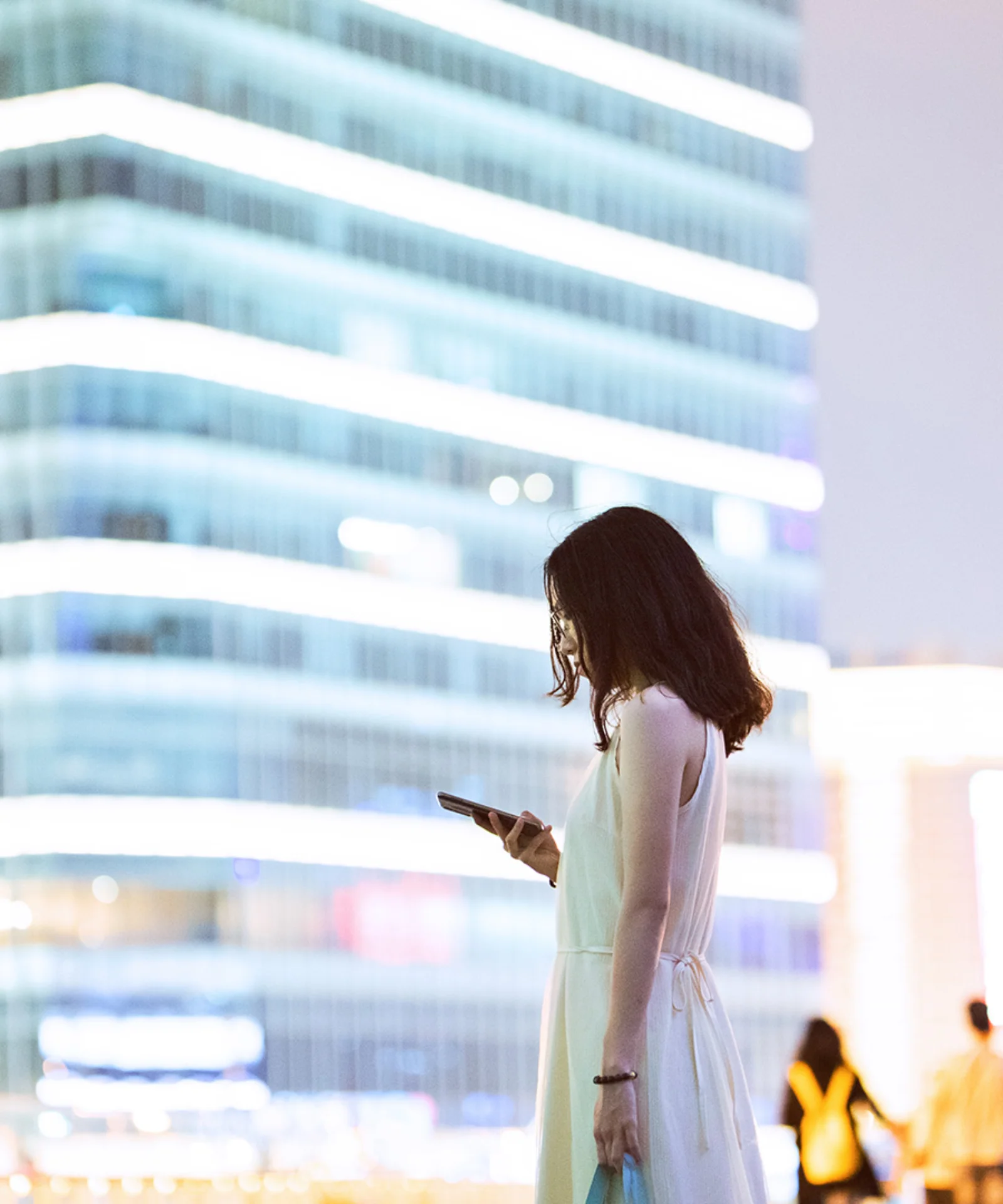 Una mujer parada en un entorno urbano por la noche, mirando su teléfono inteligente, con edificios modernos brillantemente iluminados en el fondo, que representan el tema de la digitalización en la banca.