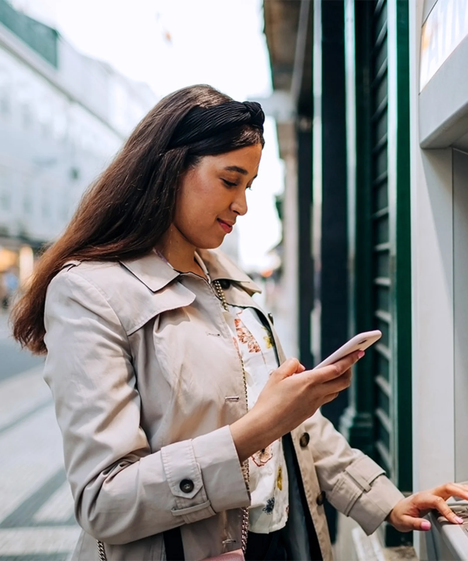 Young woman standing at an ATM while using her mobile phone, showcasing digital banking technology in an urban setting.