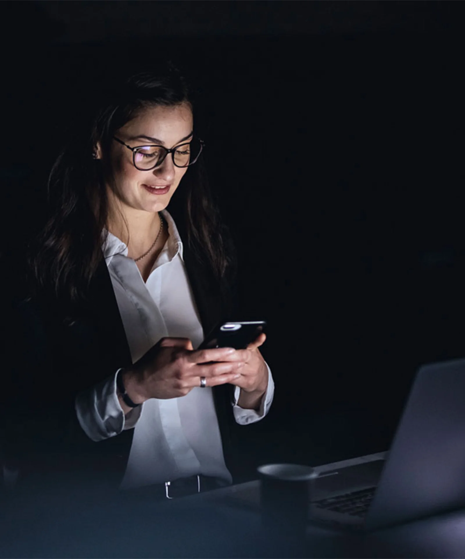 A businesswoman working late, using her smartphone while smiling, with a laptop in front of her in a dimly lit office environment.