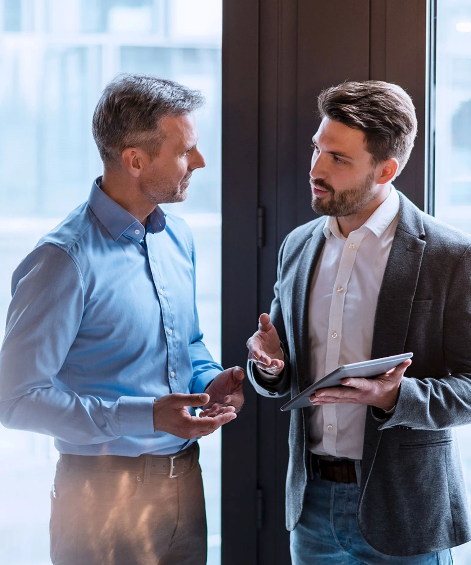 Two businessmen engaged in a discussion, one holding a tablet, in a modern office with large windows and greenery.