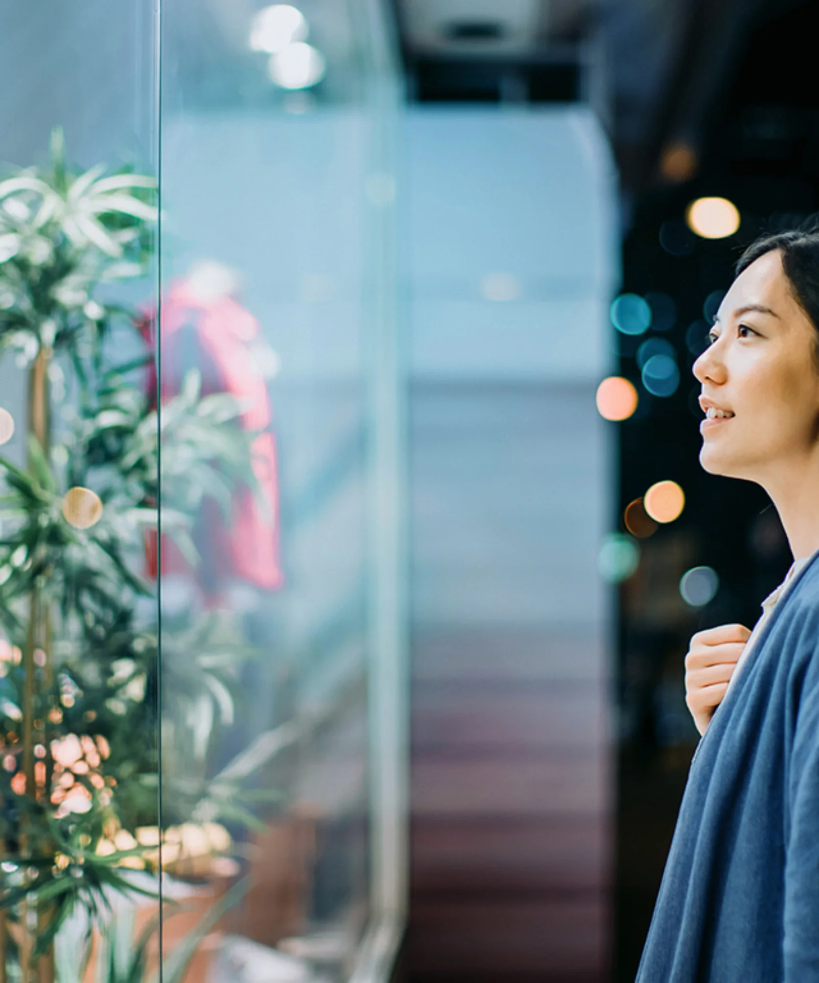 Une femme regarde la vitrine d&#039;un magasin la nuit, son reflet étant visible sur la vitre. La scène est illuminée par les lumières de la ville en arrière-plan.