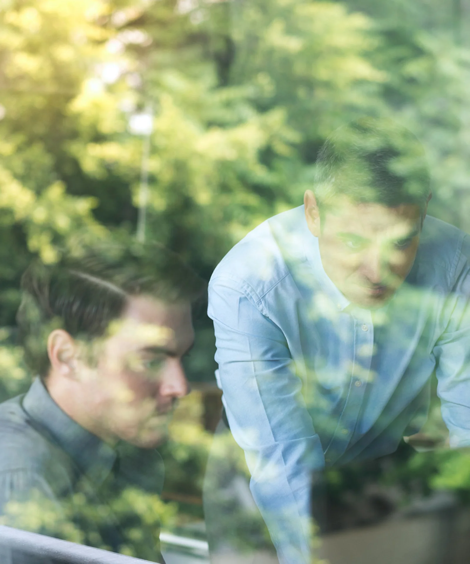 Zwei Fachleute bei der Zusammenarbeit, gesehen durch ein Fenster, in dem sich üppiges Grün spiegelt, was Zusammenarbeit und Nachhaltigkeit in der Innovation symbolisiert.