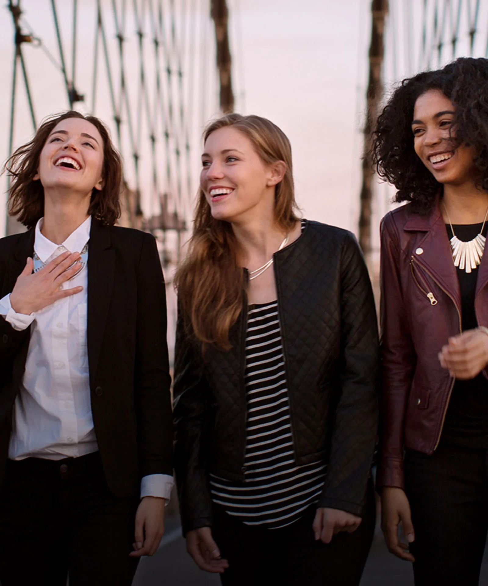 Three diverse female professionals walking and laughing together on a city bridge, exemplifying GFT&#039;s commitment to teamwork and innovation.