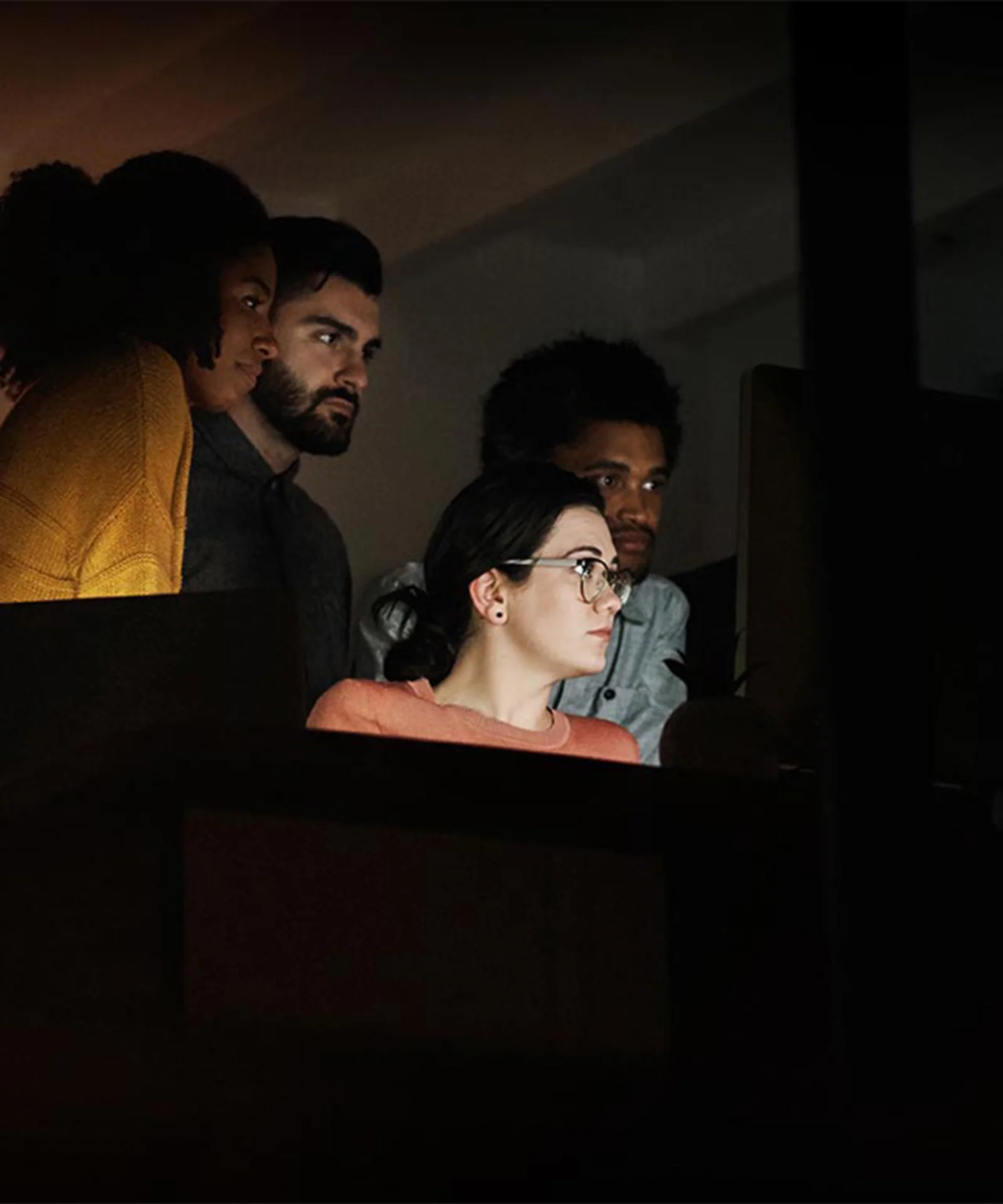 Uma equipe focada de quatro profissionais trabalhando juntos em um projeto tarde da noite, iluminados pelo brilho das telas de computador em um escritório mal iluminado.