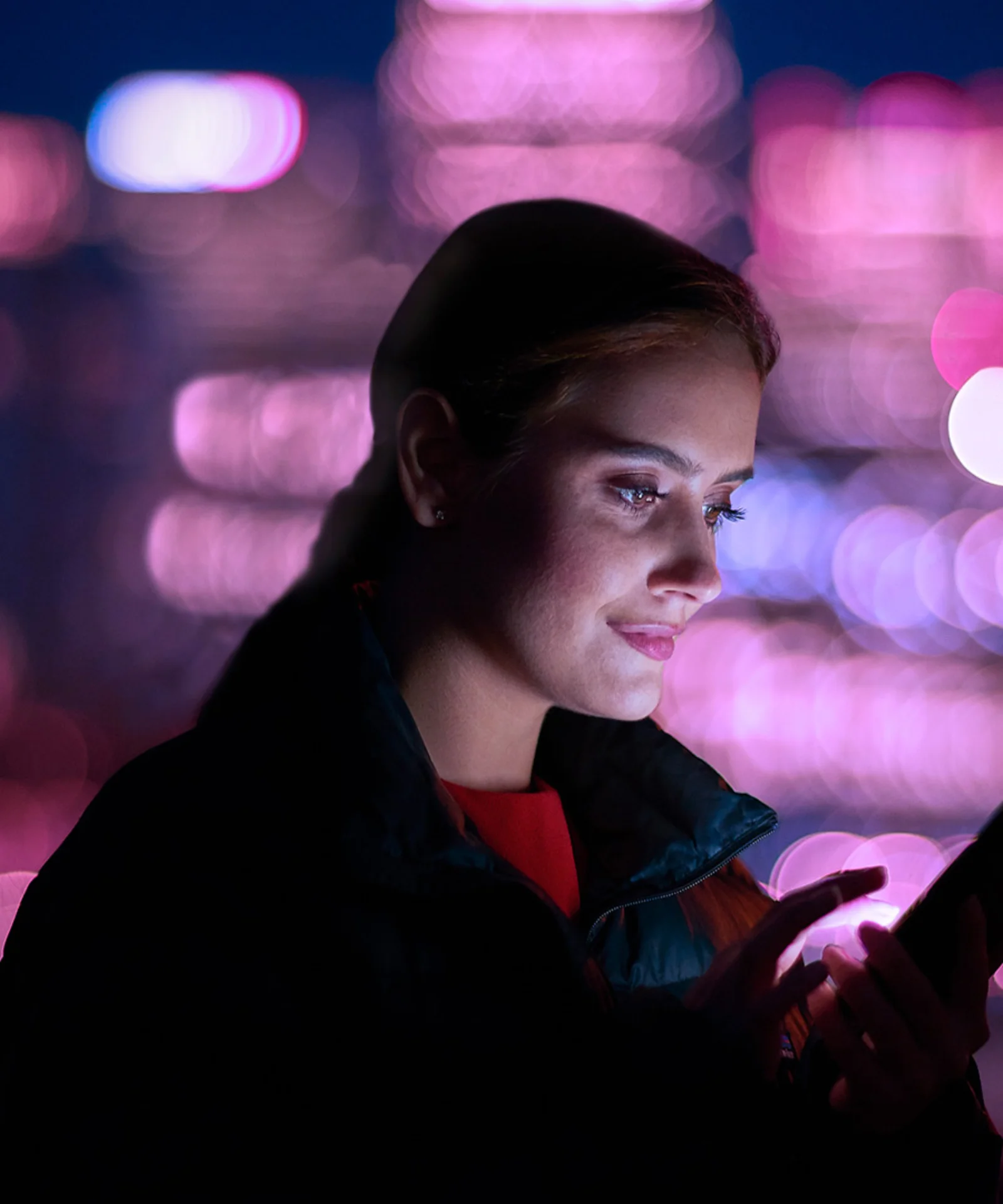 A woman using a smartphone against a backdrop of a cityscape illuminated by pink and purple lights, symbolising the transition of Guidewire to cloud-based solutions.