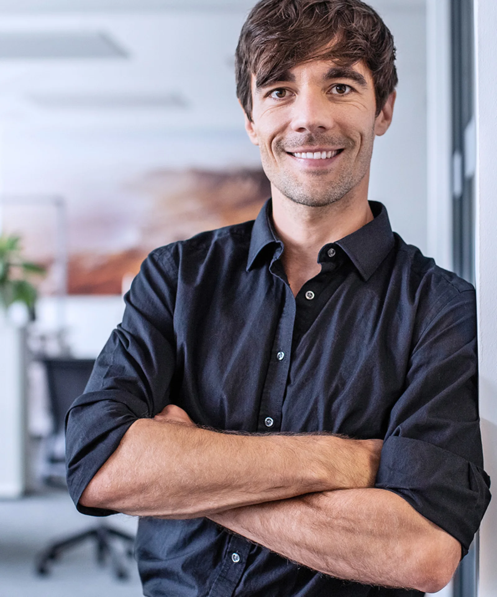 Professional man in a black shirt standing with arms crossed in an office environment.