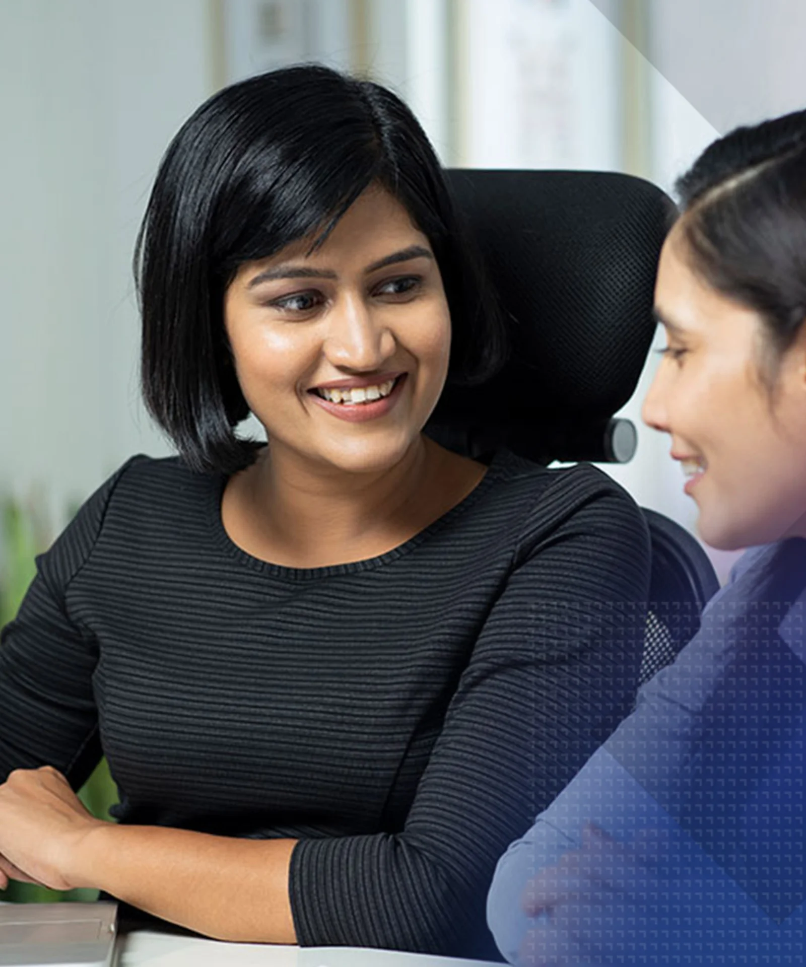 Two professional women engaged in a discussion at a desk, with a laptop open and one holding a pen, in a modern office environment.