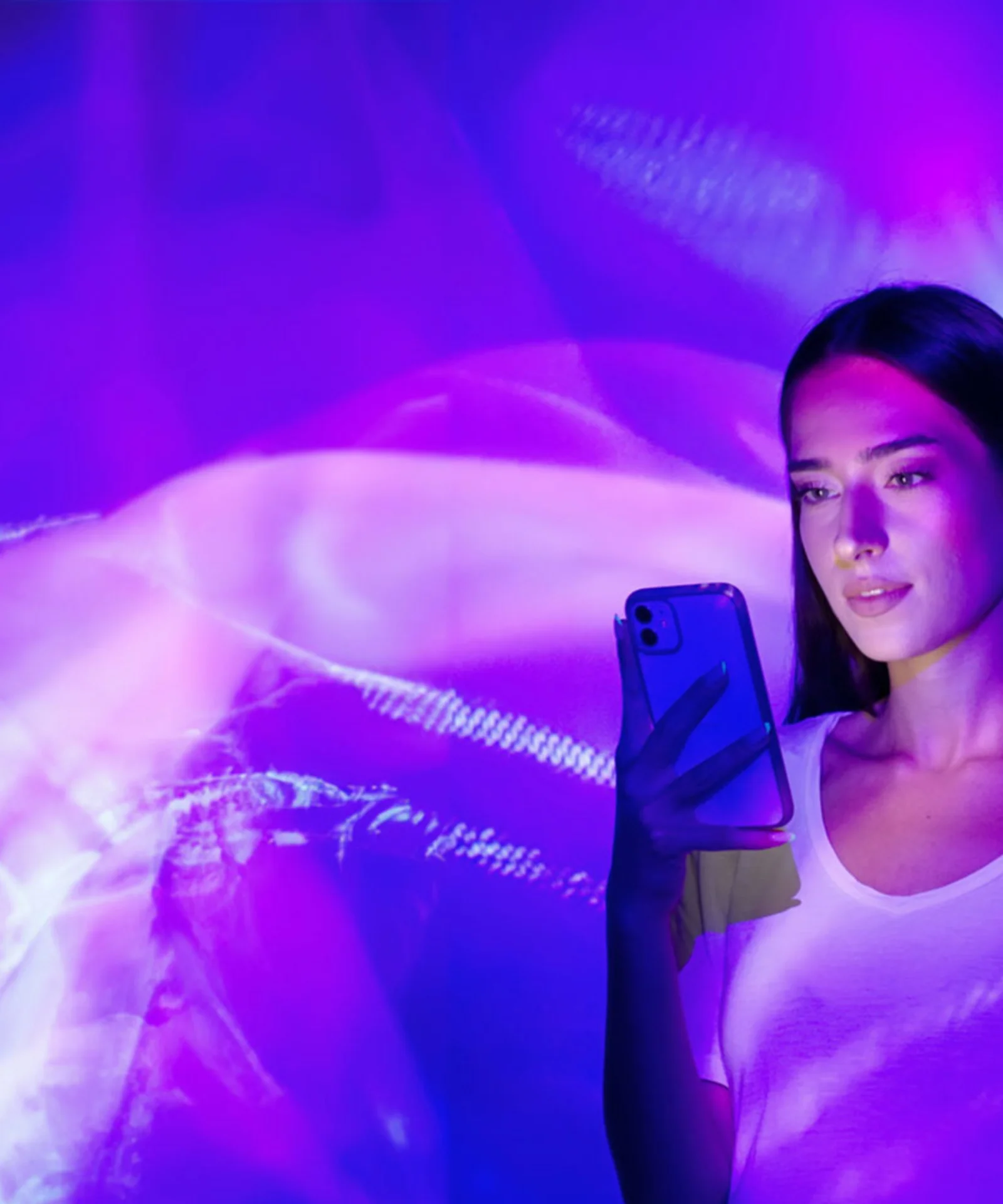 Una mujer que sostiene un teléfono inteligente se encuentra frente a una pantalla de luz vibrante y abstracta de color púrpura y rosa.