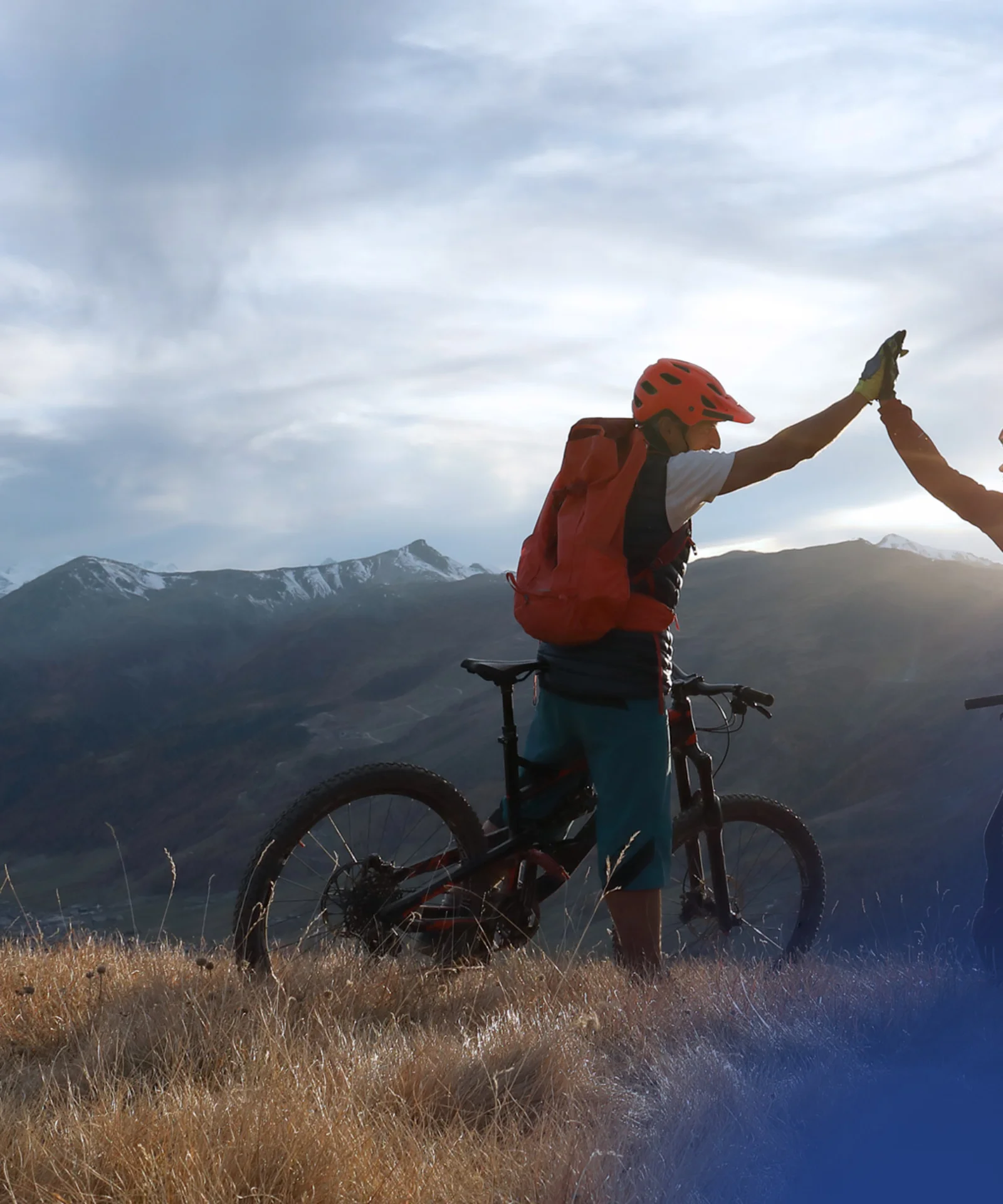 Two cyclists giving a high-five on a mountaintop, with a stunning landscape of mountains and valleys in the background, symbolising teamwork and shared achievement.