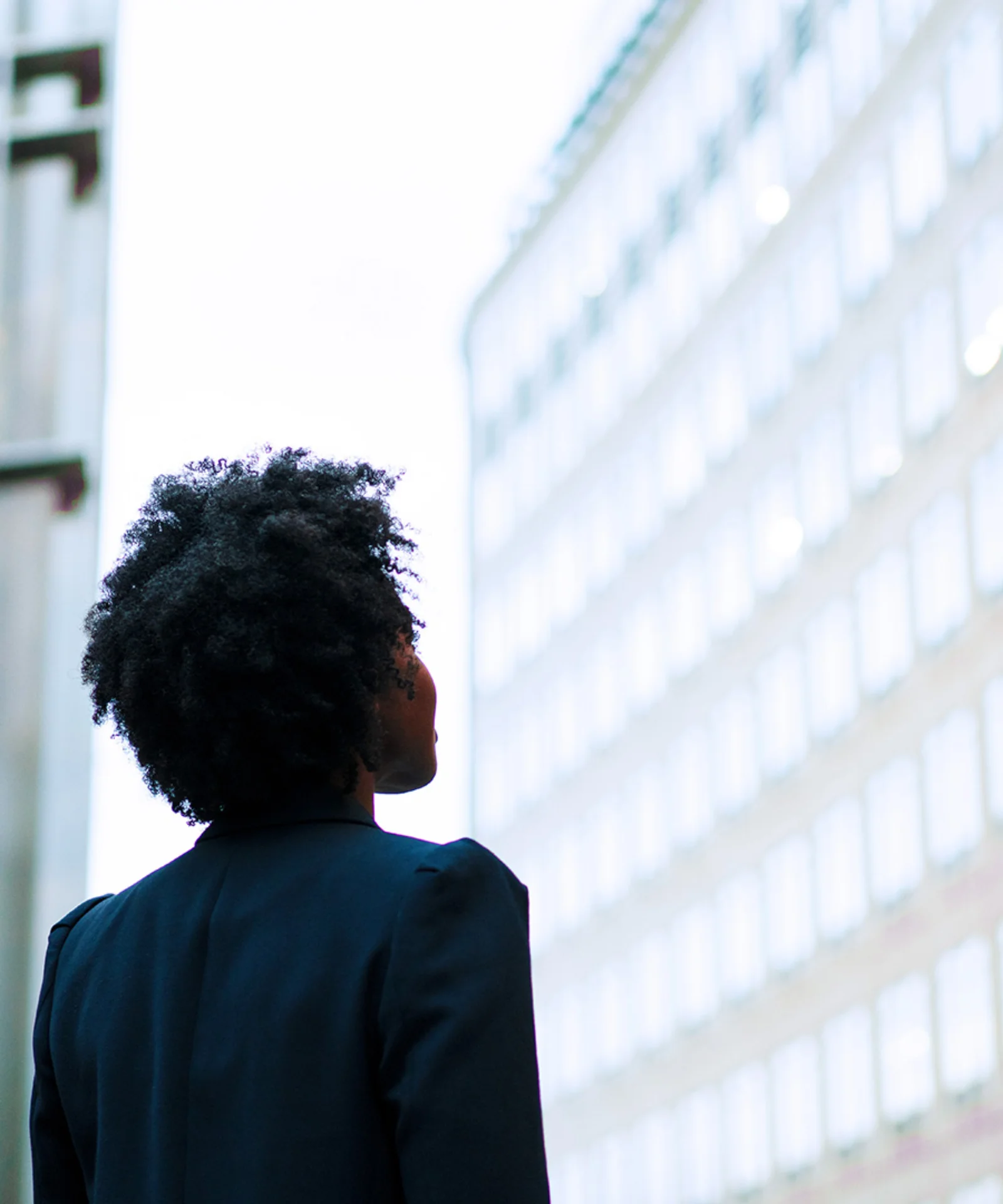 A person with natural hair, wearing a dark blazer, gazing up at modern office buildings with illuminated windows in a city environment.