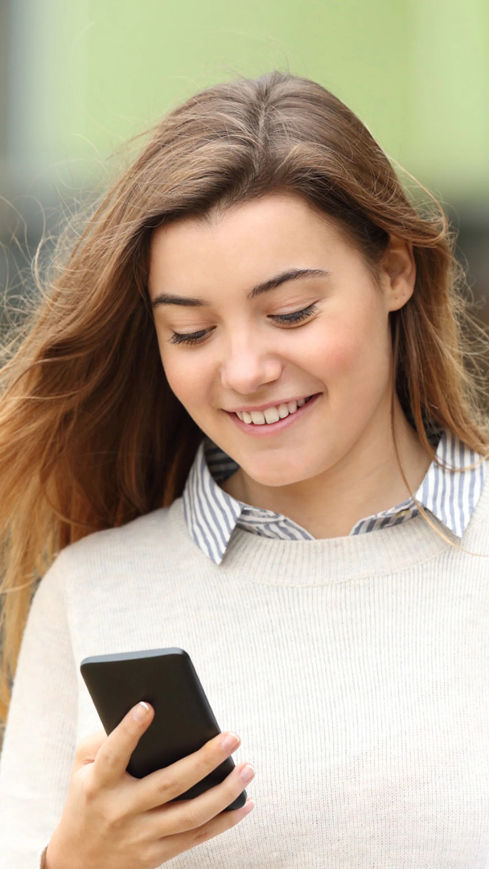 A young woman smiling while using her smartphone, holding a folder, symbolising the convenience and accessibility of the Crédit Agricole BankMeApp.