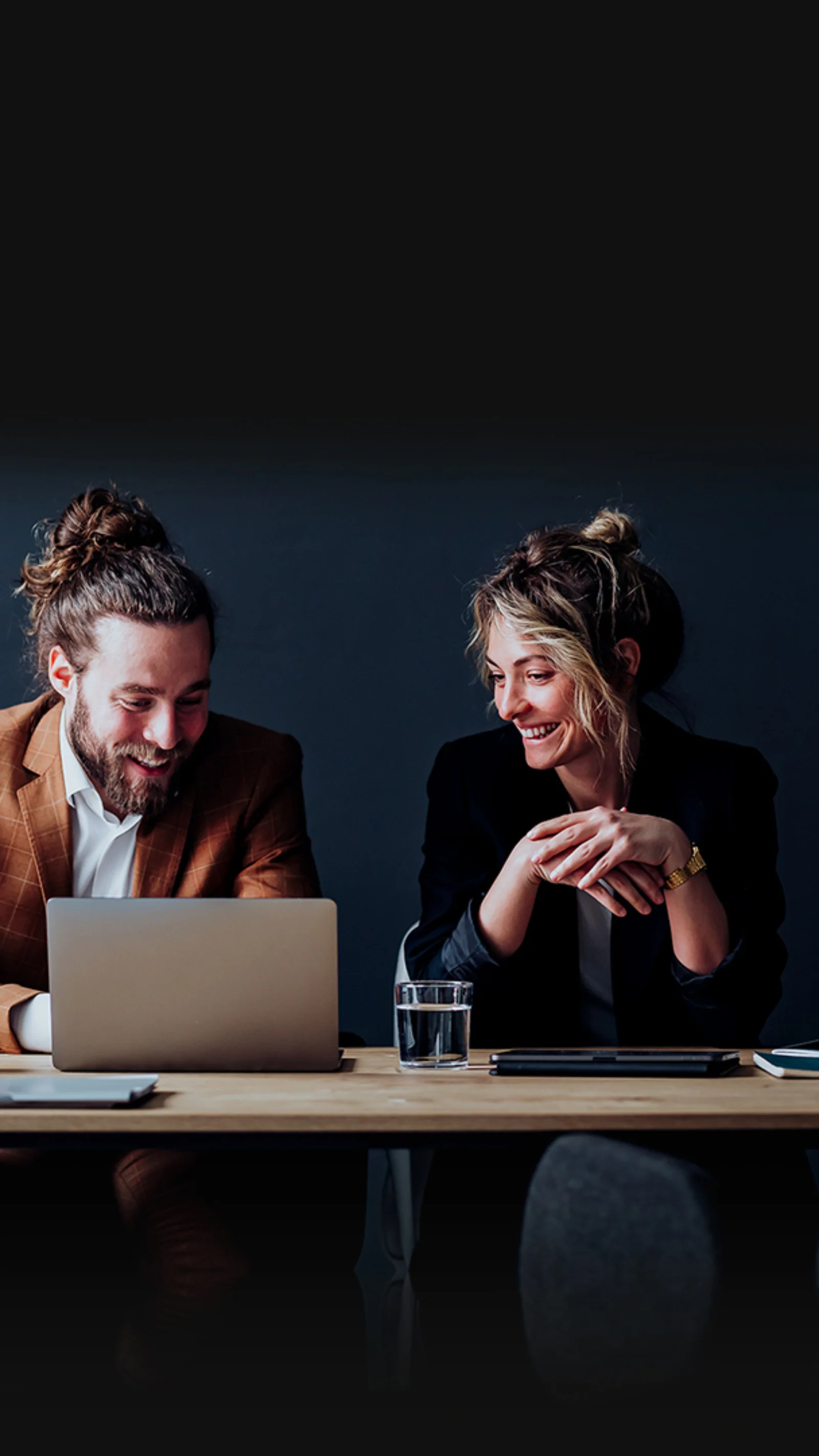 Two professionals engaged in a collaborative discussion, sitting at a desk with a laptop, notebooks, and coffee, showcasing an image design services setup.