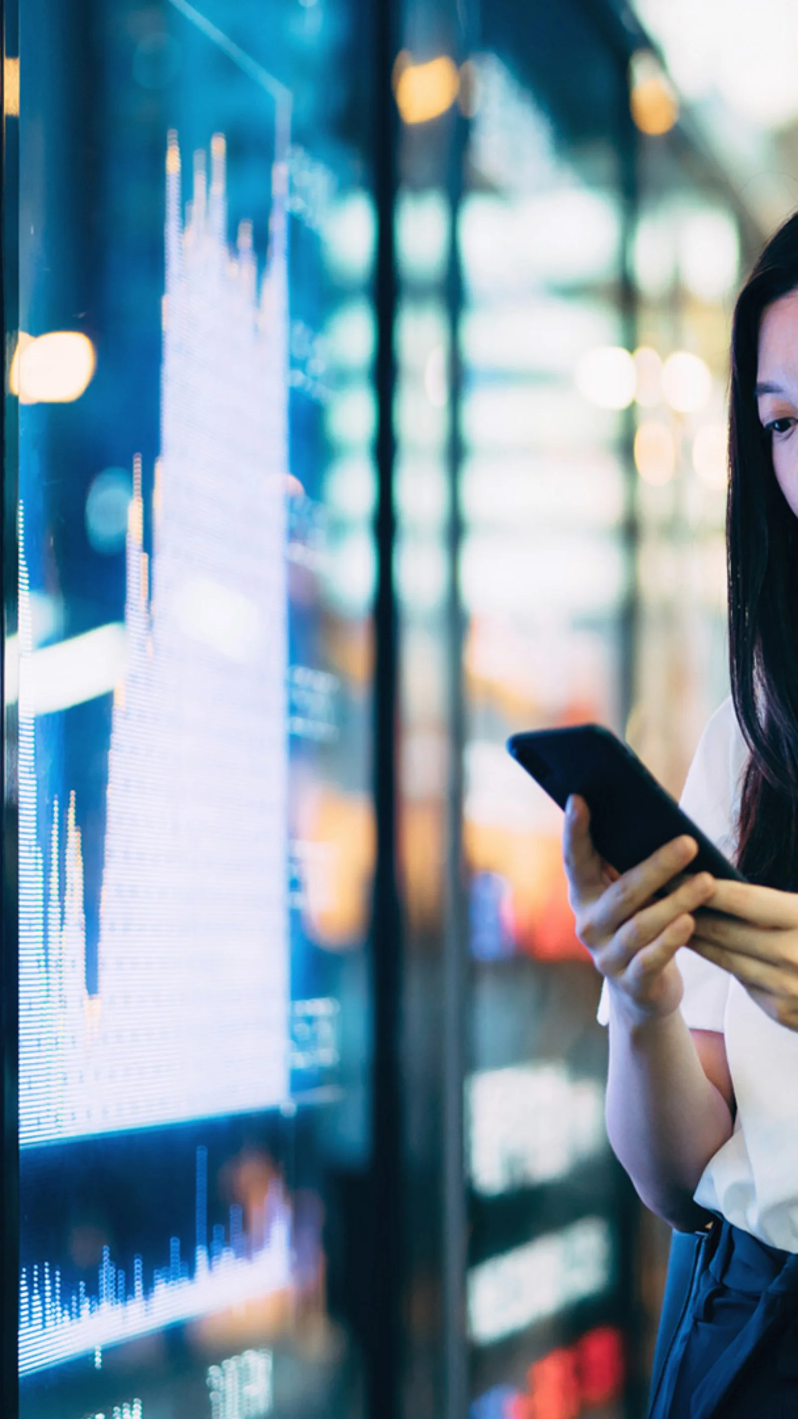 imagem mostra uma mulher usando seu smartphone enquanto está em pé na frente de um quadro eletrônico que exibe dados financeiros. Este visual simboliza a integração da sustentabilidade em tecnologia e práticas comerciais, destacando a importância do desenvolvimento sustentável na era digital moderna. O uso da tecnologia no monitoramento e promoção de iniciativas sustentáveis é um tema-chave representado nesta imagem.