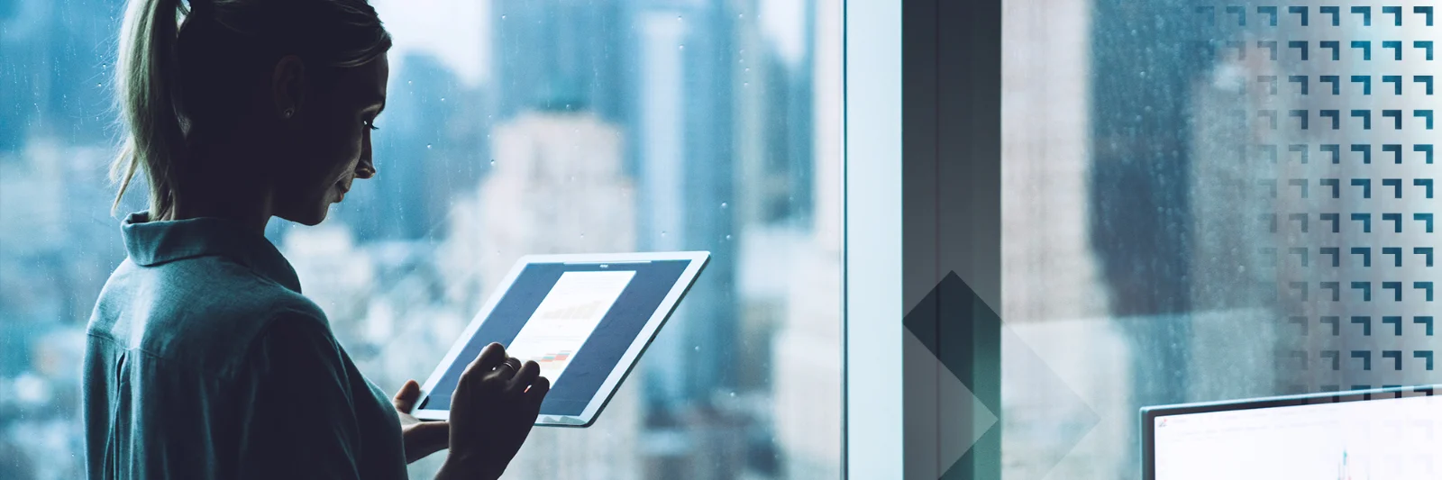 Silhouette of a professional woman analyzing data on a tablet while standing near a window overlooking a cityscape.