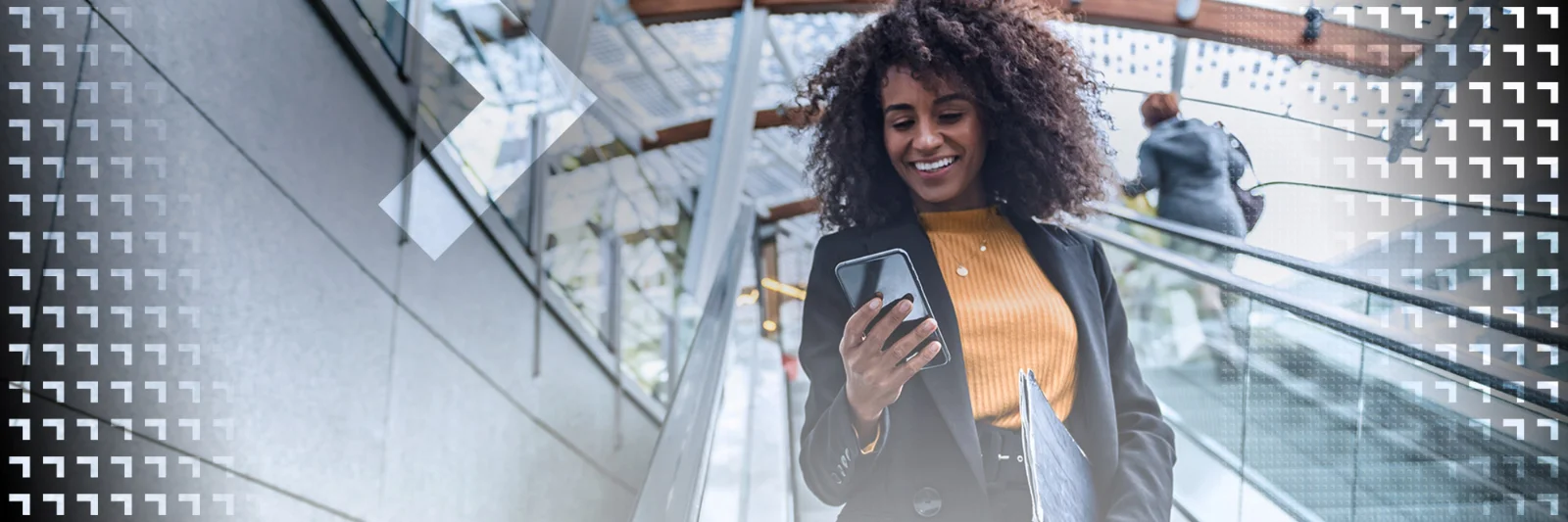 Smiling businesswoman using a smartphone while riding an escalator in a modern building with glass and metal architecture.