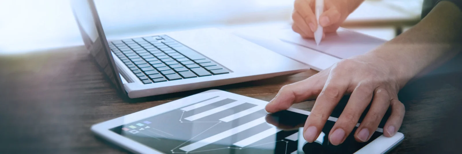 Close-up of a person working on a tablet with a laptop nearby, representing a modern digital work environment with multitasking and data analysis.