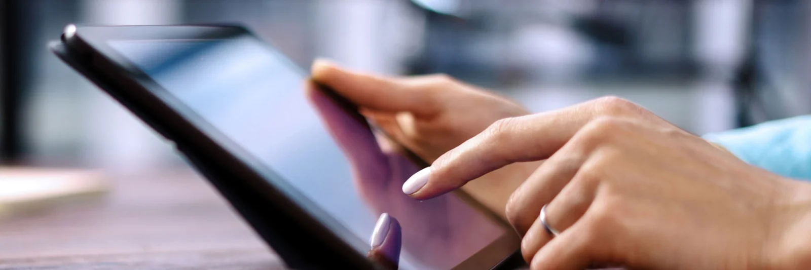 A imagem mostra um close-up de mãos usando um tablet, com uma mão tocando na tela. O tablet está sobre uma superfície e o fundo está levemente desfocado, sugerindo um espaço interior. O foco está na interação com o tablet.