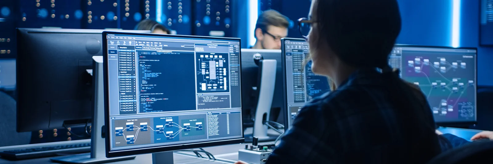 Software engineer working on multiple monitors displaying complex code and data visualisations in a high-tech office environment.