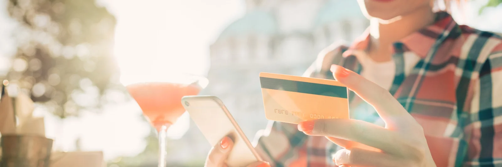 Una mujer sonriente sosteniendo un teléfono inteligente y una tarjeta de crédito, disfrutando de una bebida al aire libre, simbolizando la conveniencia y la facilidad de la banca personal moderna.