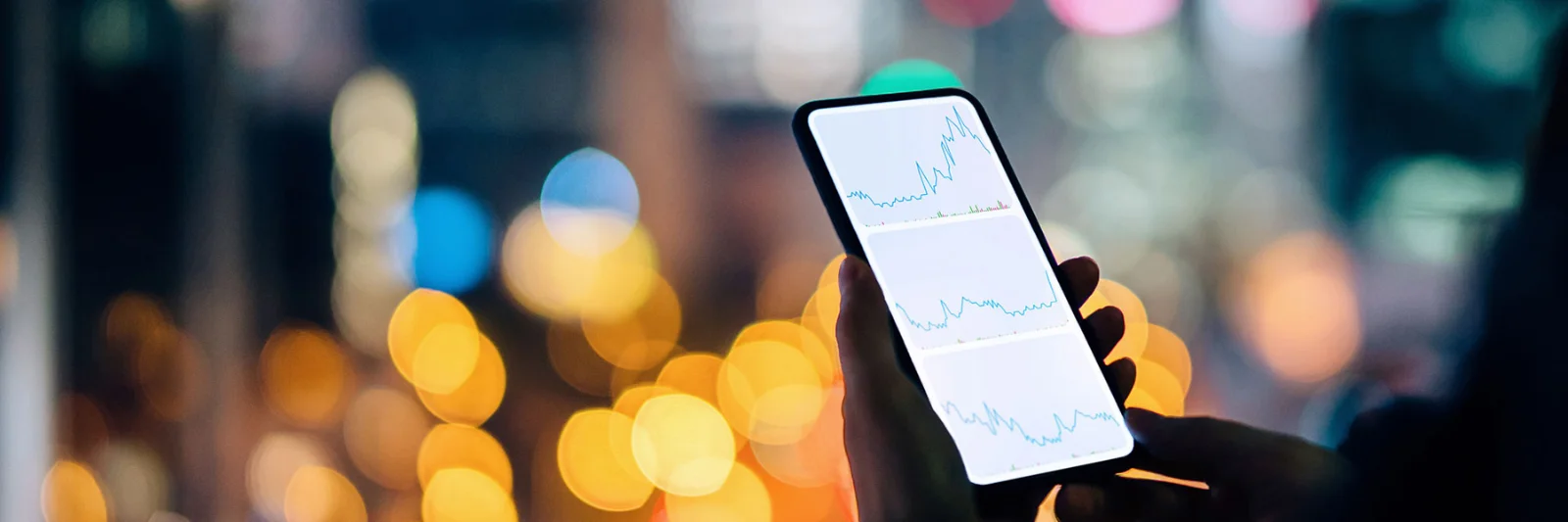 A person holding a smartphone displaying financial graphs, with a cityscape illuminated by vibrant lights in the background.