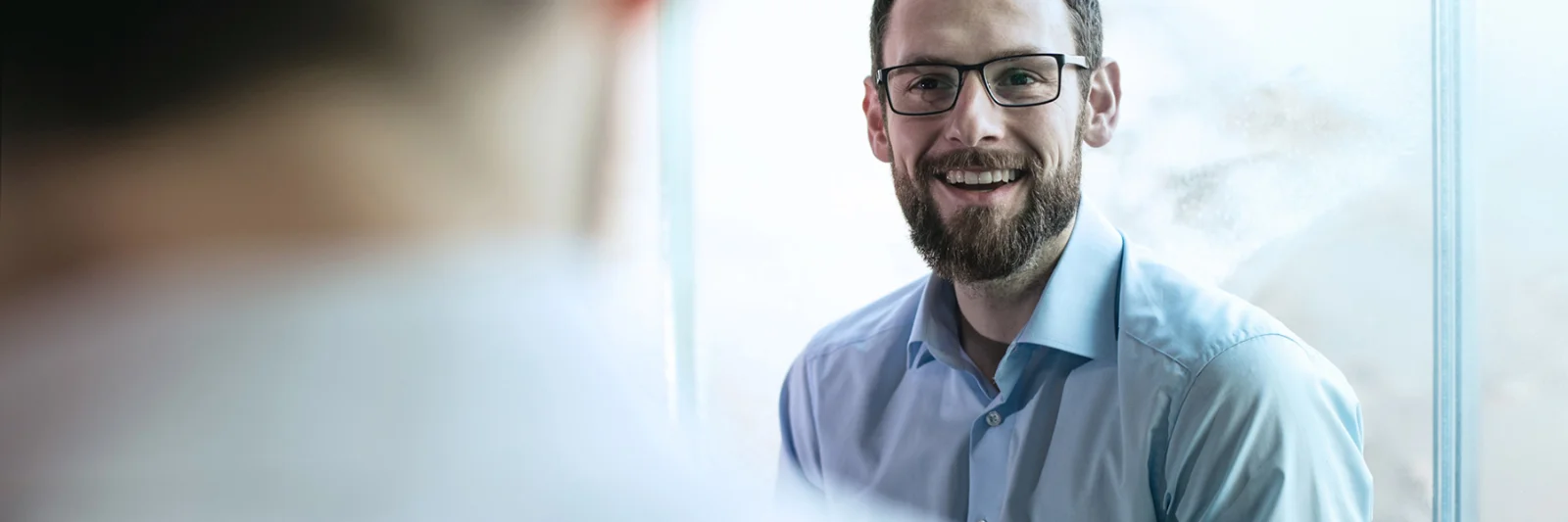 A business professional with glasses smiling during a conversation in a bright office setting