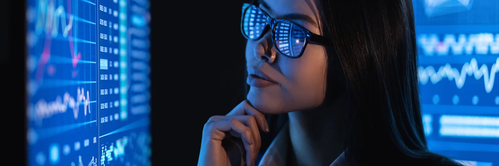 Una mujer con gafas, reflejando gráficos de datos, mira atentamente varias pantallas digitales que muestran diversos análisis y gráficos.