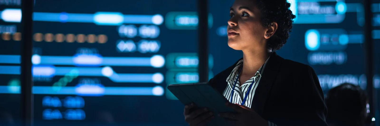 An IT professional using a tablet while overseeing digital operations in a high-tech control room with multiple data displays.