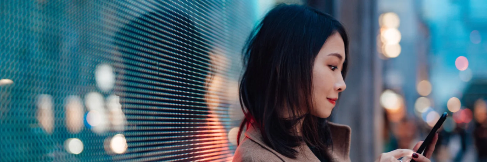 A woman standing against a digital screen at night, using her smartphone. The screen&#039;s light reflects on her face and the surrounding area, creating a modern, tech-savvy ambiance.