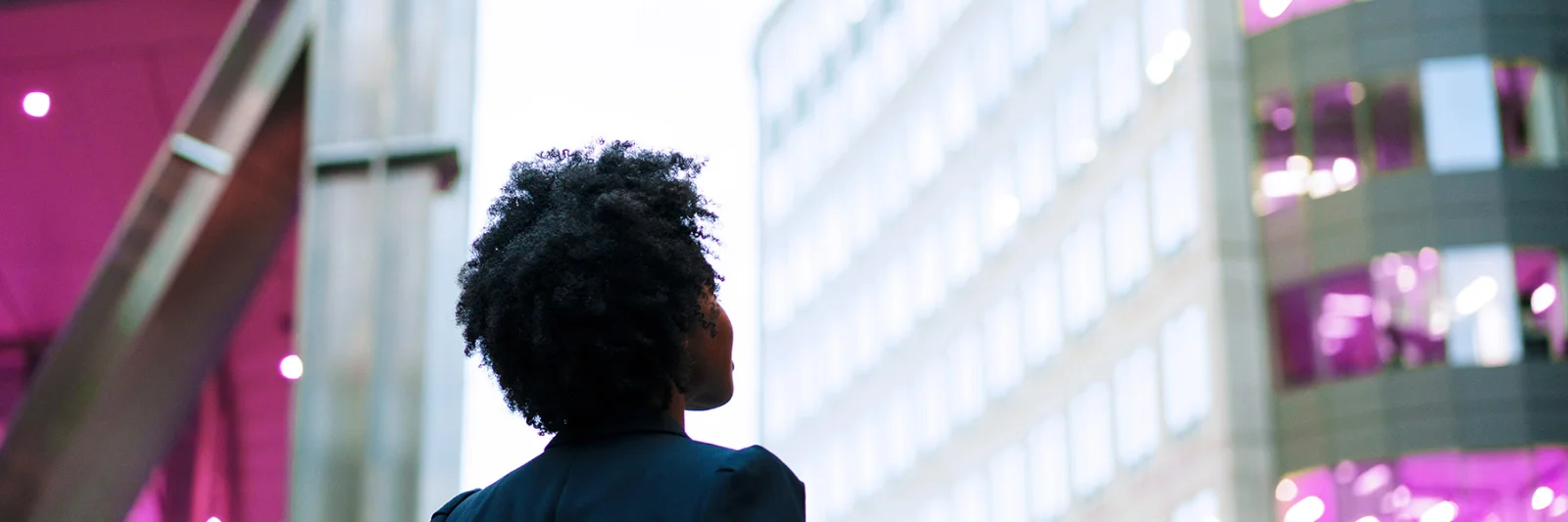 A person with natural hair, wearing a dark blazer, gazing up at modern office buildings with illuminated windows in a city environment.