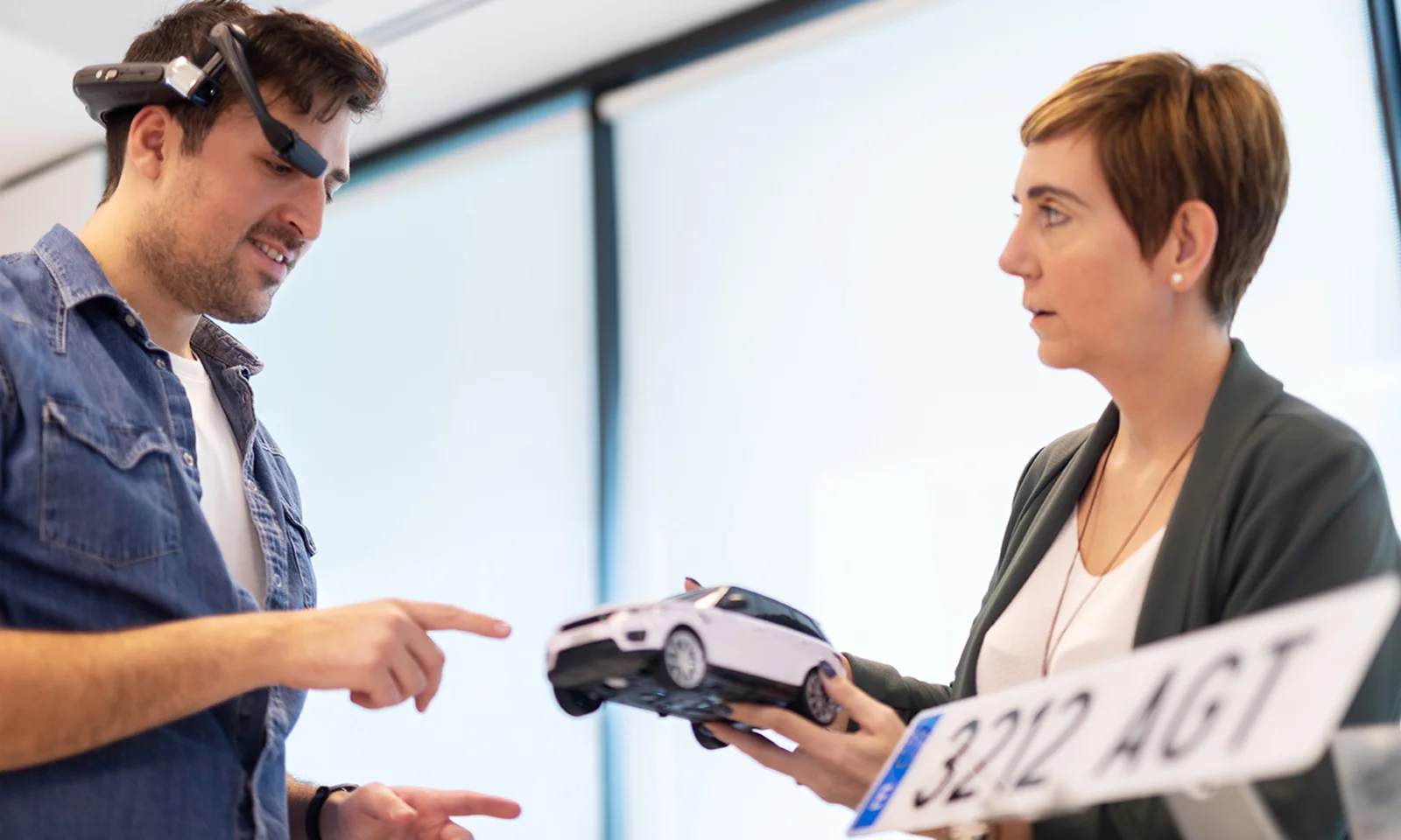 Two professionals collaborating in a design product innovation workshop. One person is wearing an augmented reality headset and pointing, while the other holds a model car and a license plate.