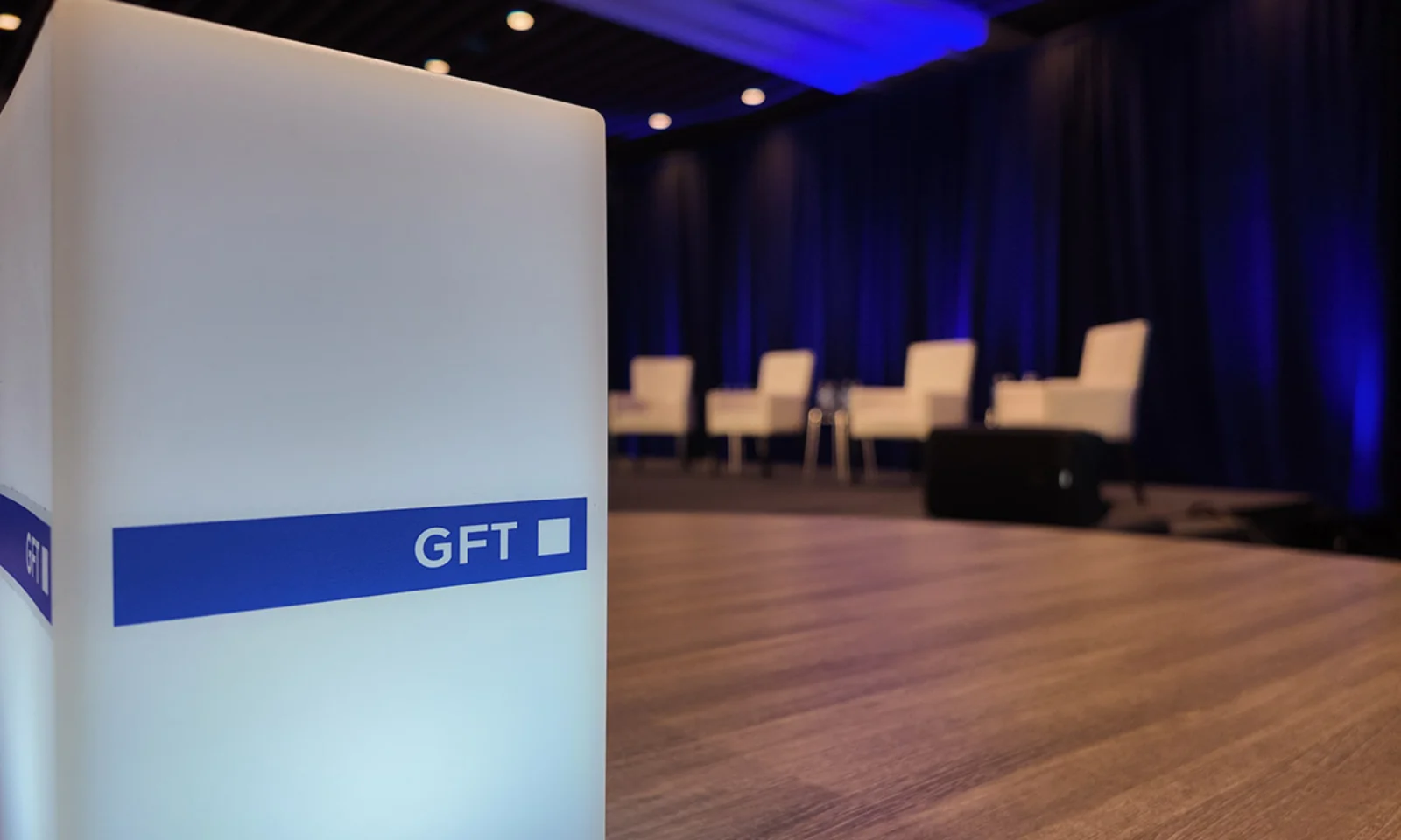 Close-up of a glowing cube with the GFT logo in the foreground, against a blurred stage setup with white chairs and a blue-lit backdrop.