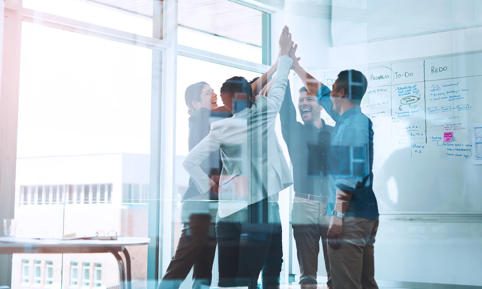 A group of professionals high-fiving in an office setting, celebrating a successful collaboration with whiteboards and brainstorming notes in the background.