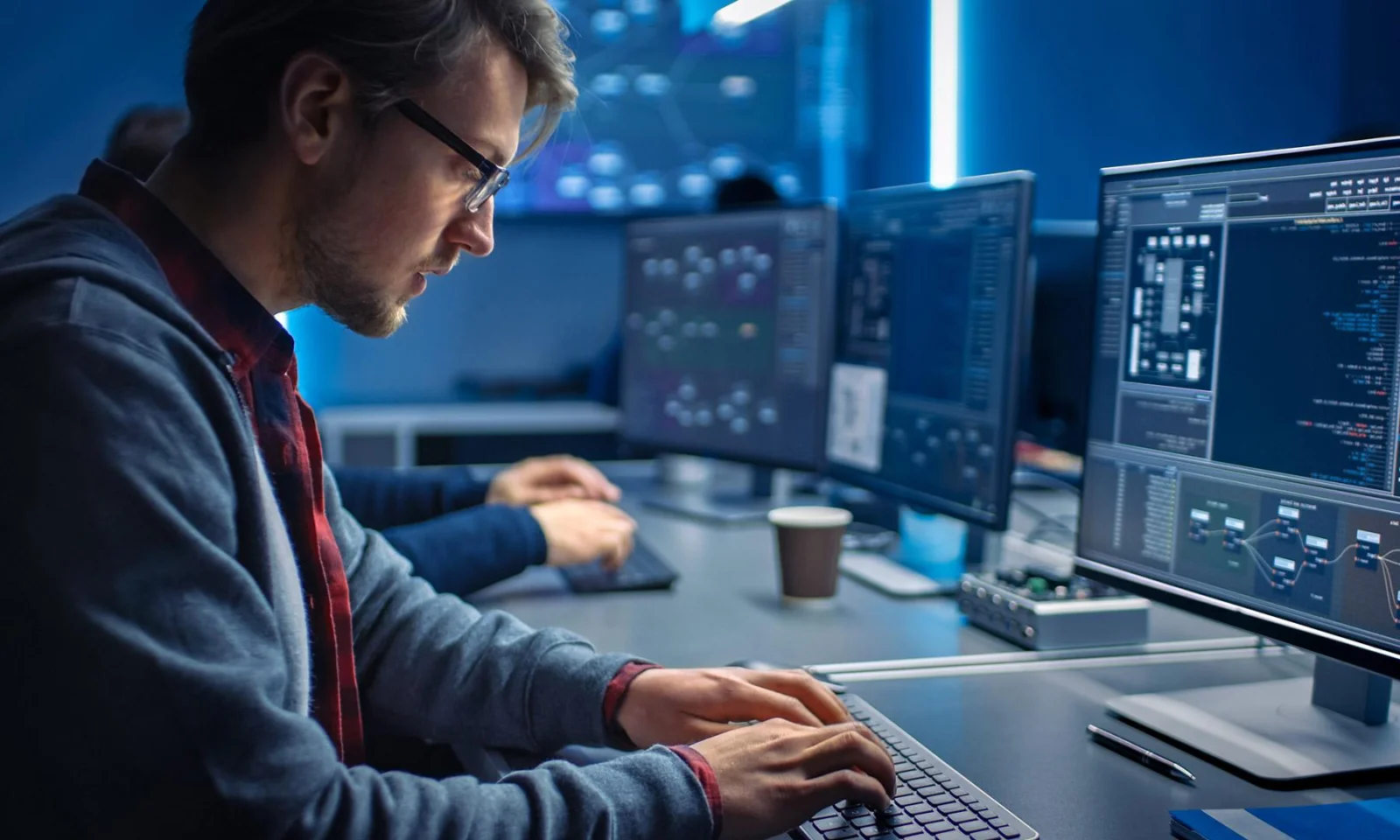 A focused software developer wearing glasses and a hoodie works on a computer with multiple screens displaying code and data.