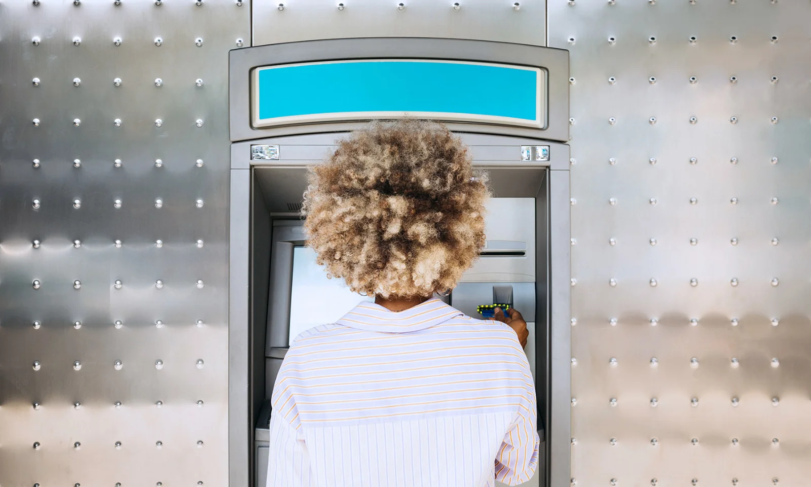 Back view of a person with curly hair using an ATM, inserting a card for a financial transaction.