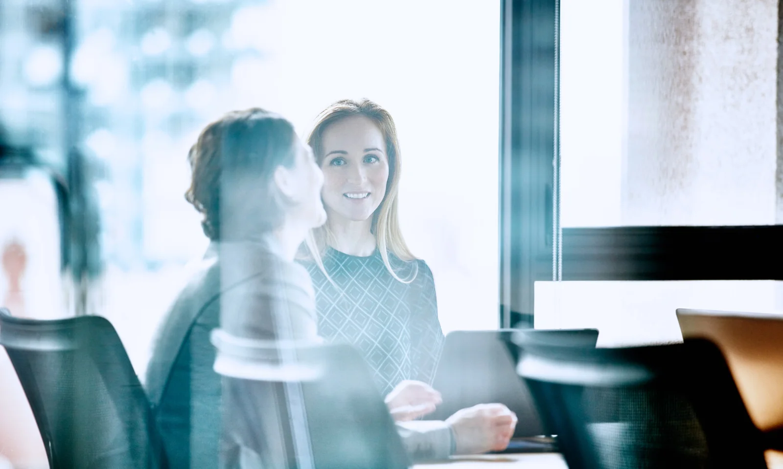 Two professional women engaged in a conversation during a business meeting, captured through a glass window, highlighting collaboration and workplace dynamics.