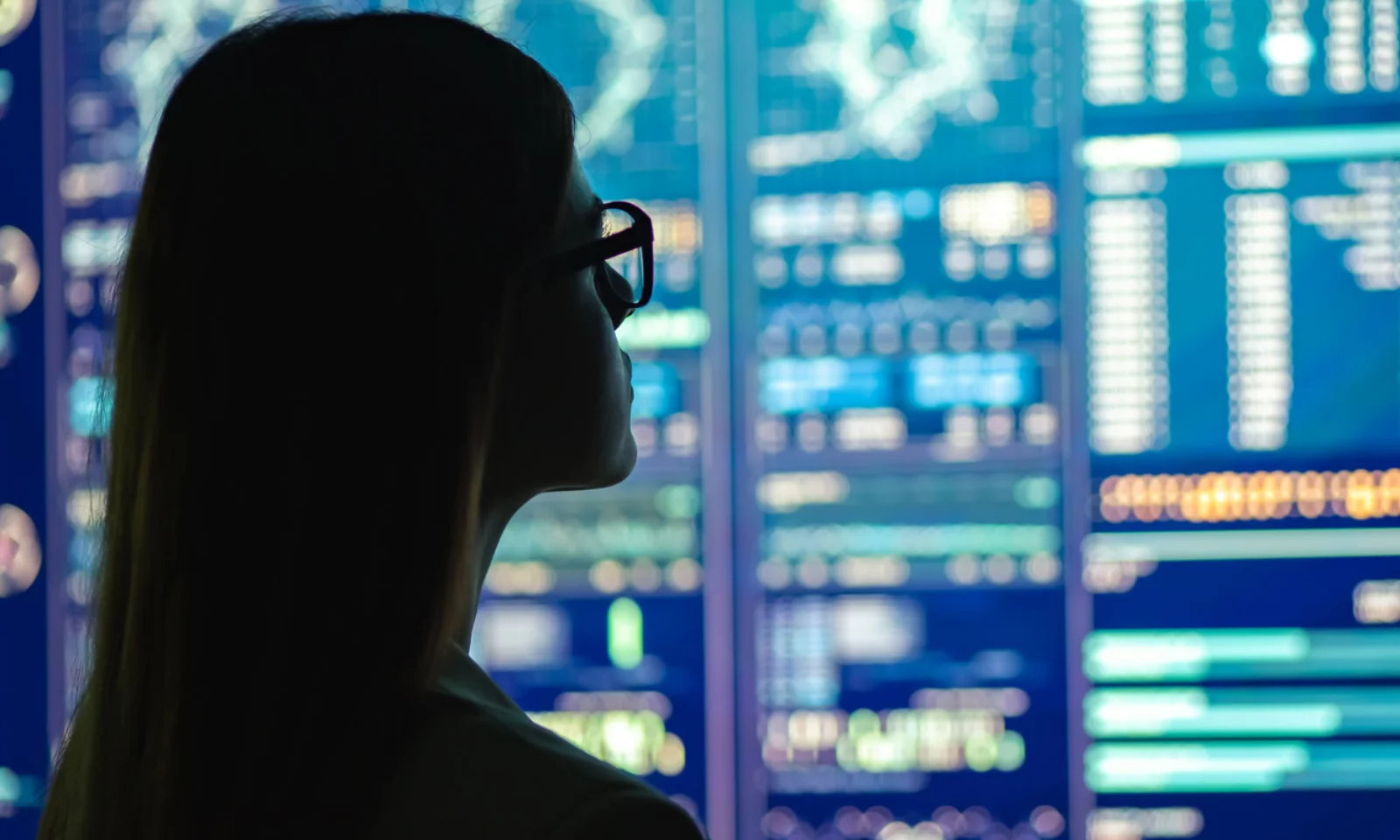 Silhouette of a professional woman wearing glasses, analyzing complex data displayed on multiple large digital screens.