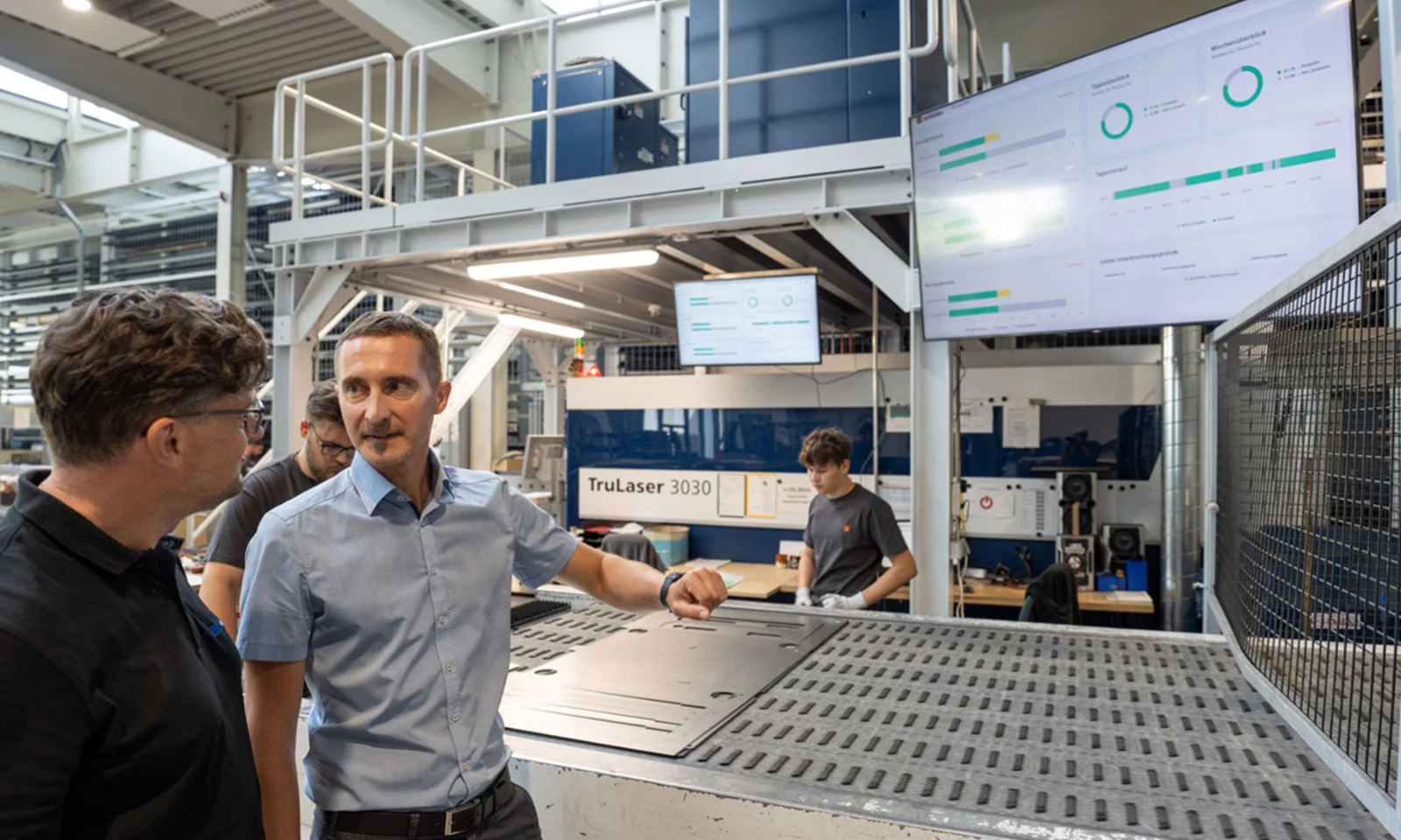 Two professionals discuss operations on the shopfloor at Schinko, with real-time data displayed on screens in the background, highlighting the transparency and efficiency in the manufacturing process.