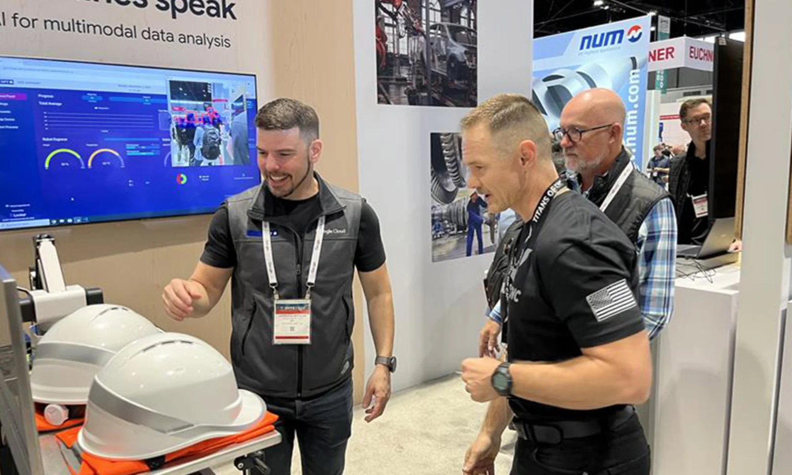 Trade show attendees interacting with an industrial data analysis display, featuring real-time performance metrics on a screen and protective helmets being examined for safety testing.