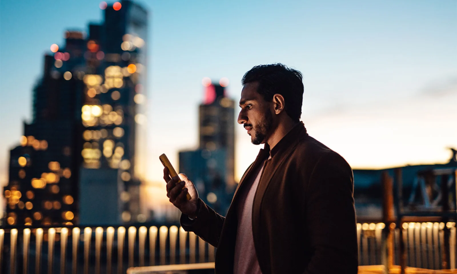 A professional man in a brown coat using a smartphone against the backdrop of a city skyline illuminated by evening lights.