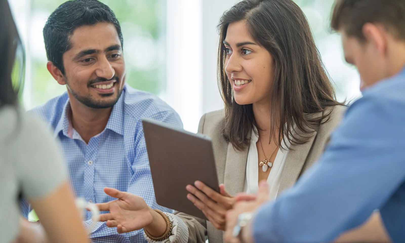 A group of professionals engaged in a collaborative discussion, with a woman holding a tablet and sharing ideas in a bright, modern office setting.