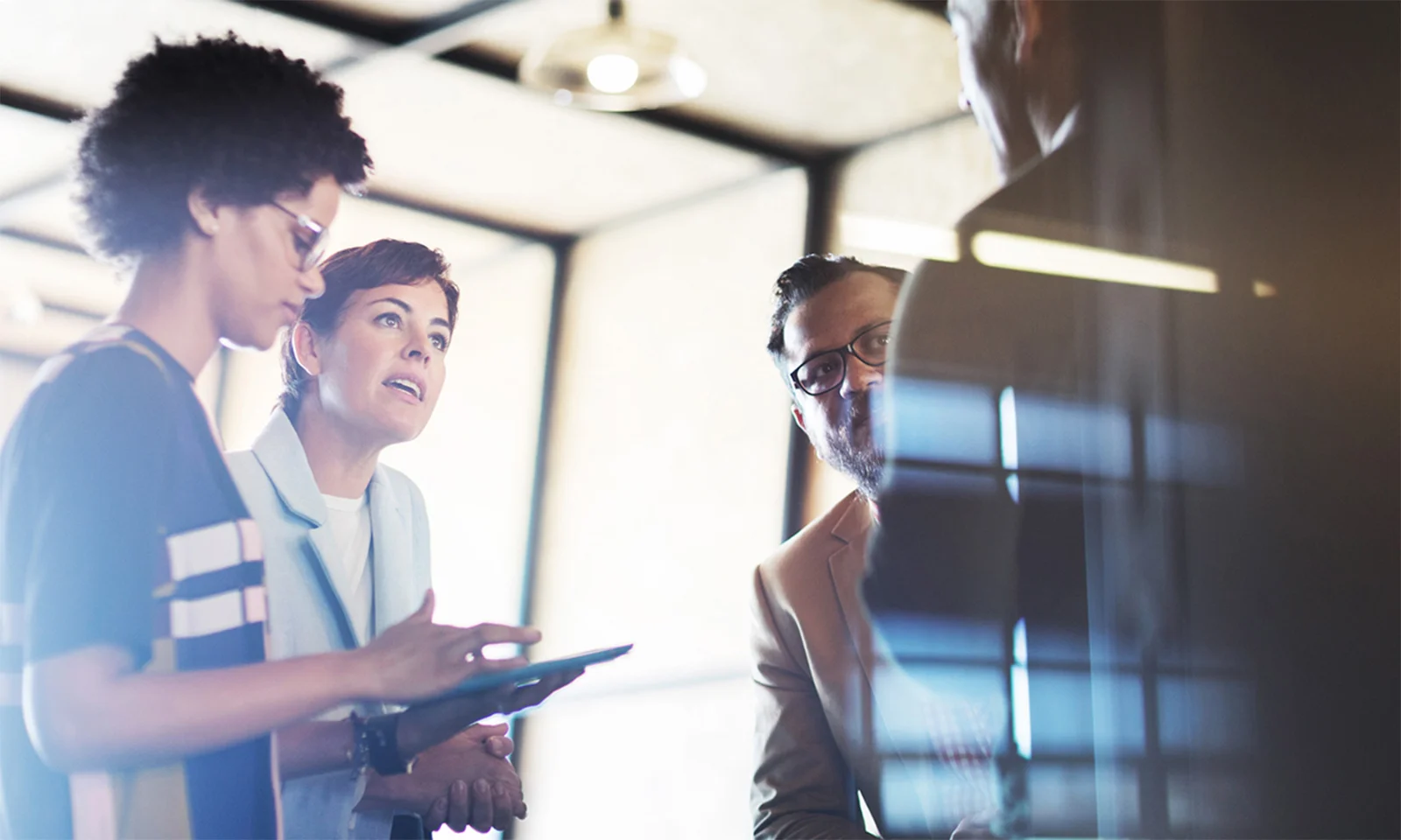 A diverse group of professionals engaged in a collaborative meeting, discussing digital solutions while using a tablet in a modern office environment.