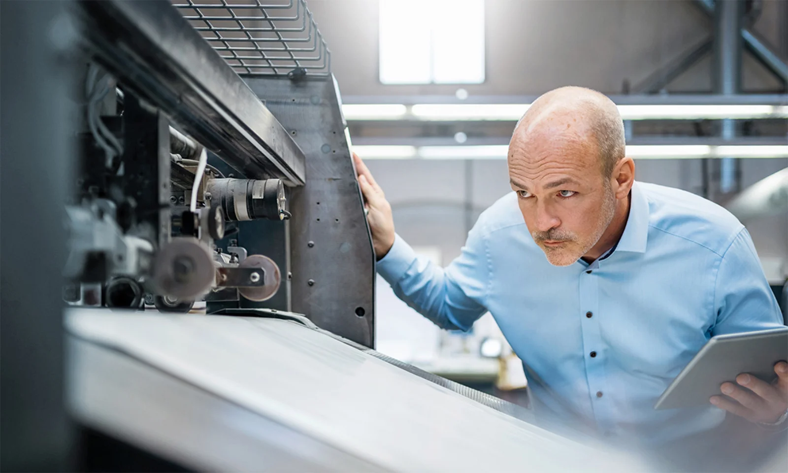 A man in a blue shirt closely inspecting machinery while holding a tablet in an industrial setting, representing quality inspection processes.