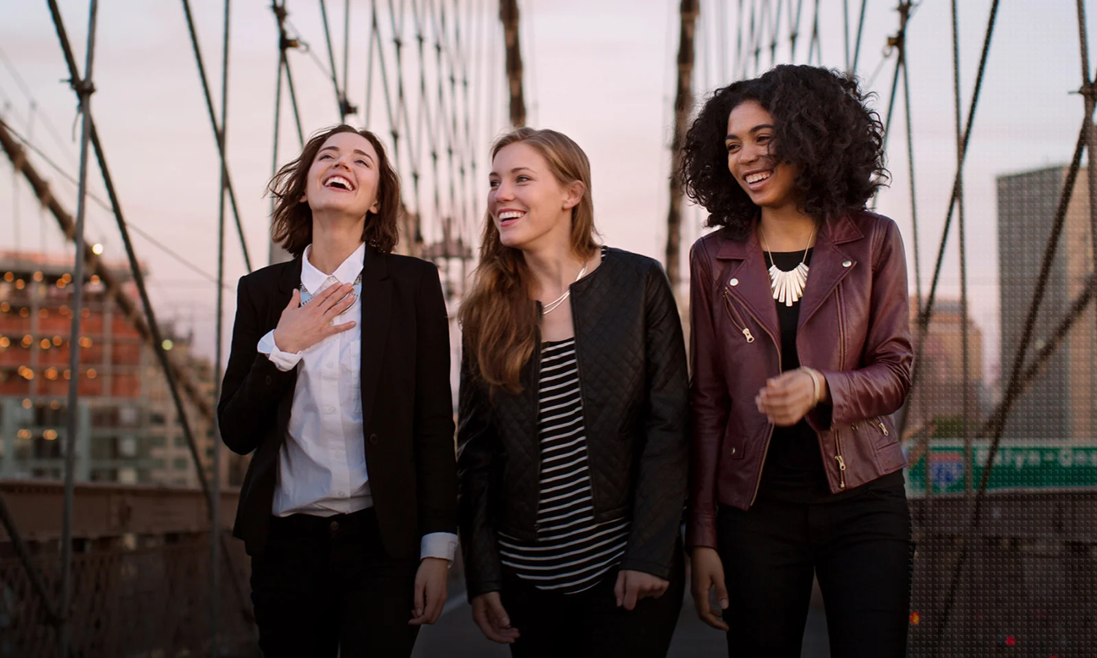 Three diverse female professionals walking and laughing together on a city bridge, exemplifying GFT&#039;s commitment to teamwork and innovation.