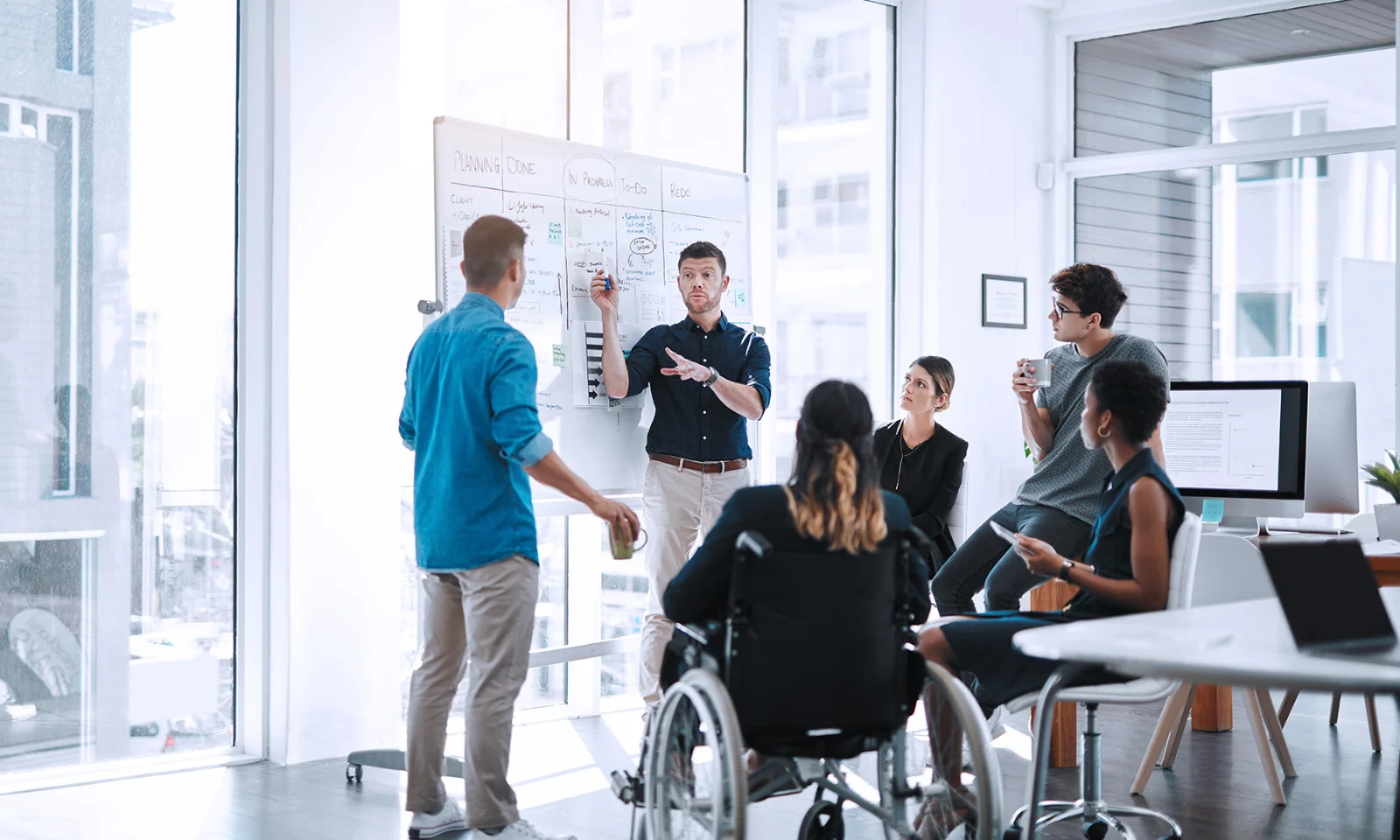 A diverse team of professionals, including a person in a wheelchair, engaging in a collaborative meeting around a whiteboard in a bright, modern office space.