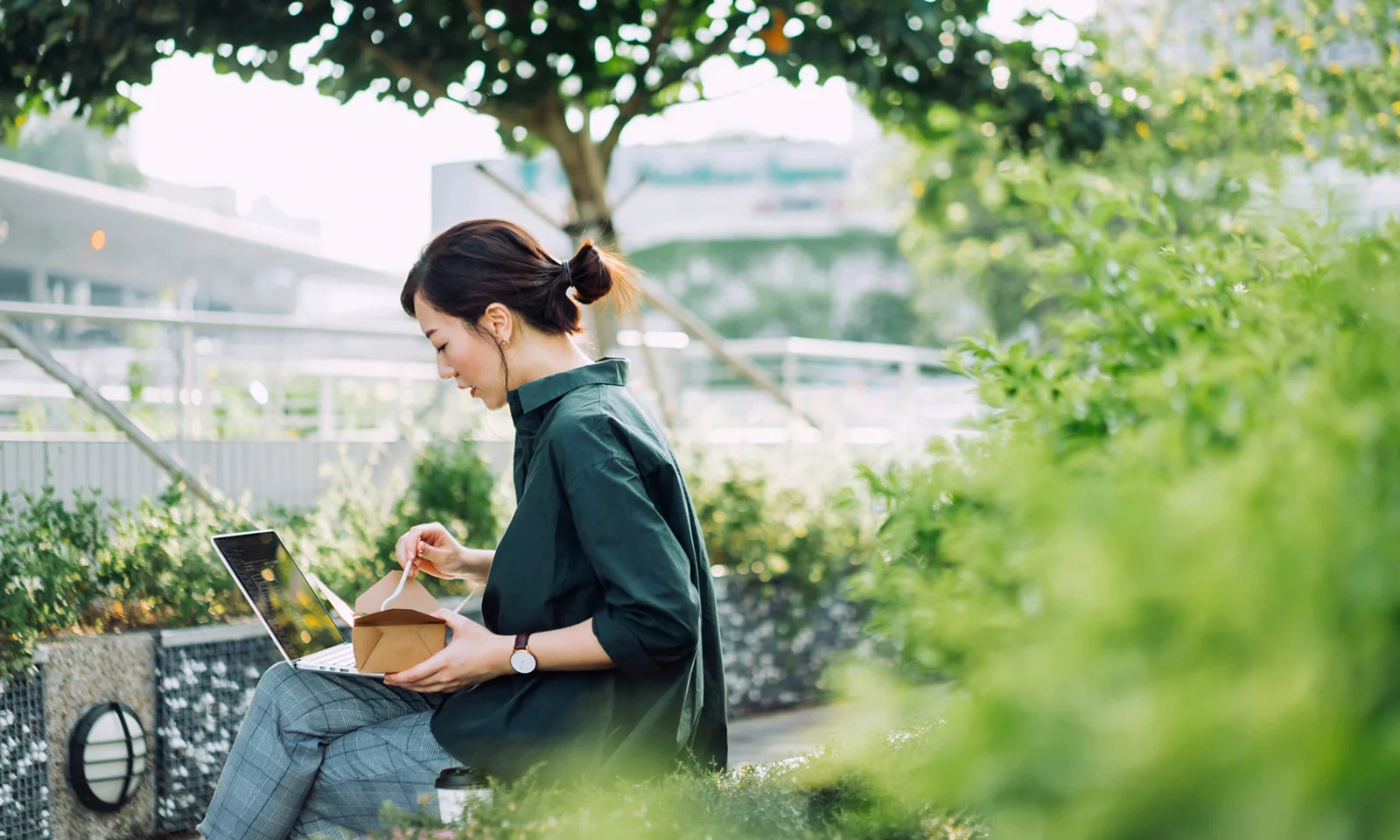 Uma mulher trabalhando em um laptop ao ar livre, cercada por vegetação, enquanto come de uma caixa de comida para viagem, simbolizando a integração de práticas verdes na vida profissional diária e na sustentabilidade.