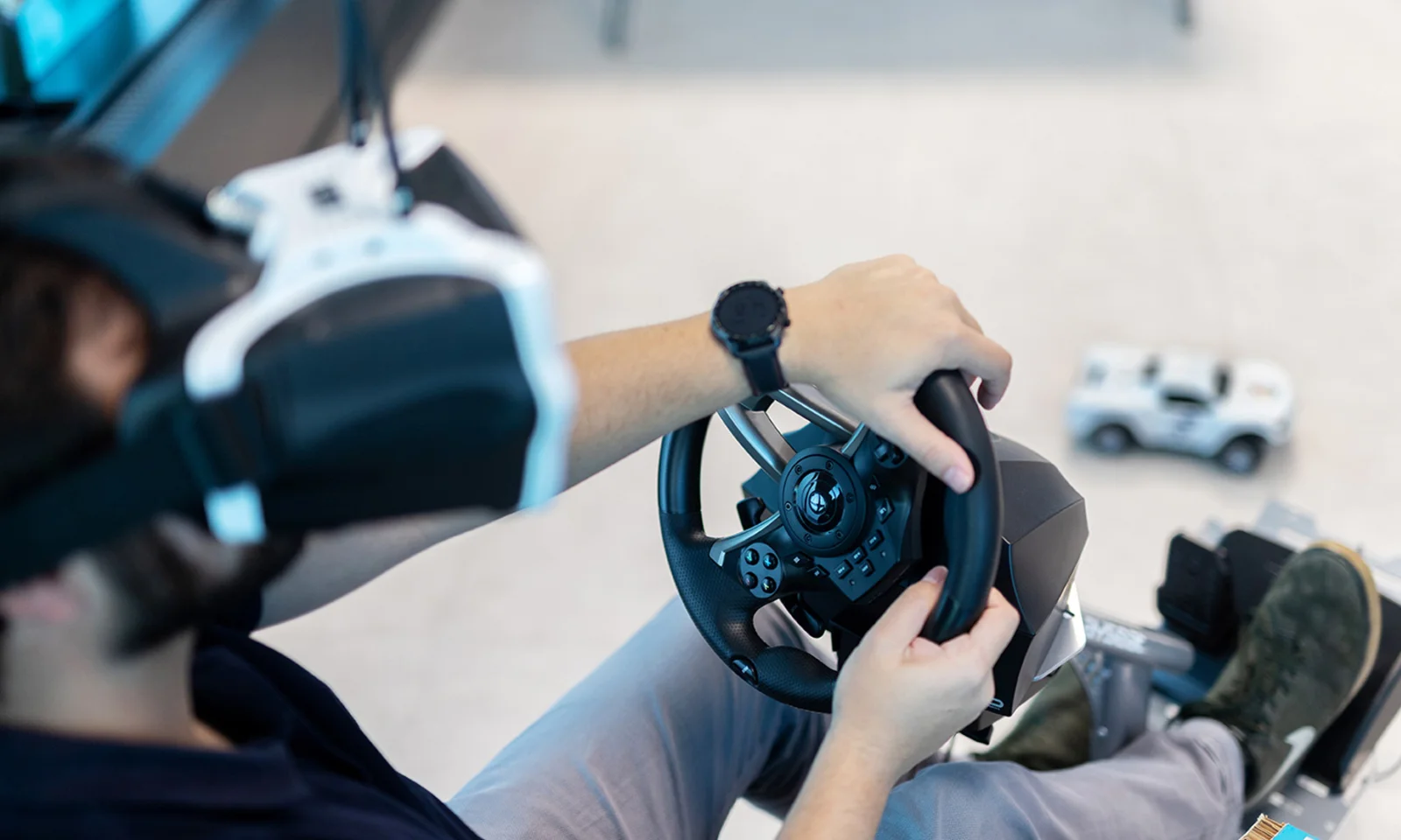 A participant in a design product innovation workshop using a virtual reality (VR) headset and steering wheel setup, simulating a driving experience. A model car is visible on the floor, indicating the immersive nature of the workshop.